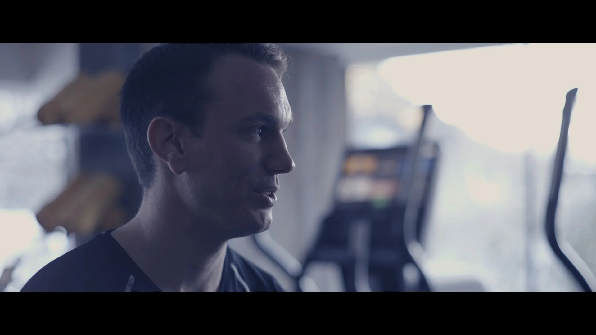 Ein Mann trainiert auf einem Heimtrainer im Fitnessstudio, das im Hintergrund mit Computern und Geräten ausgestattet ist.