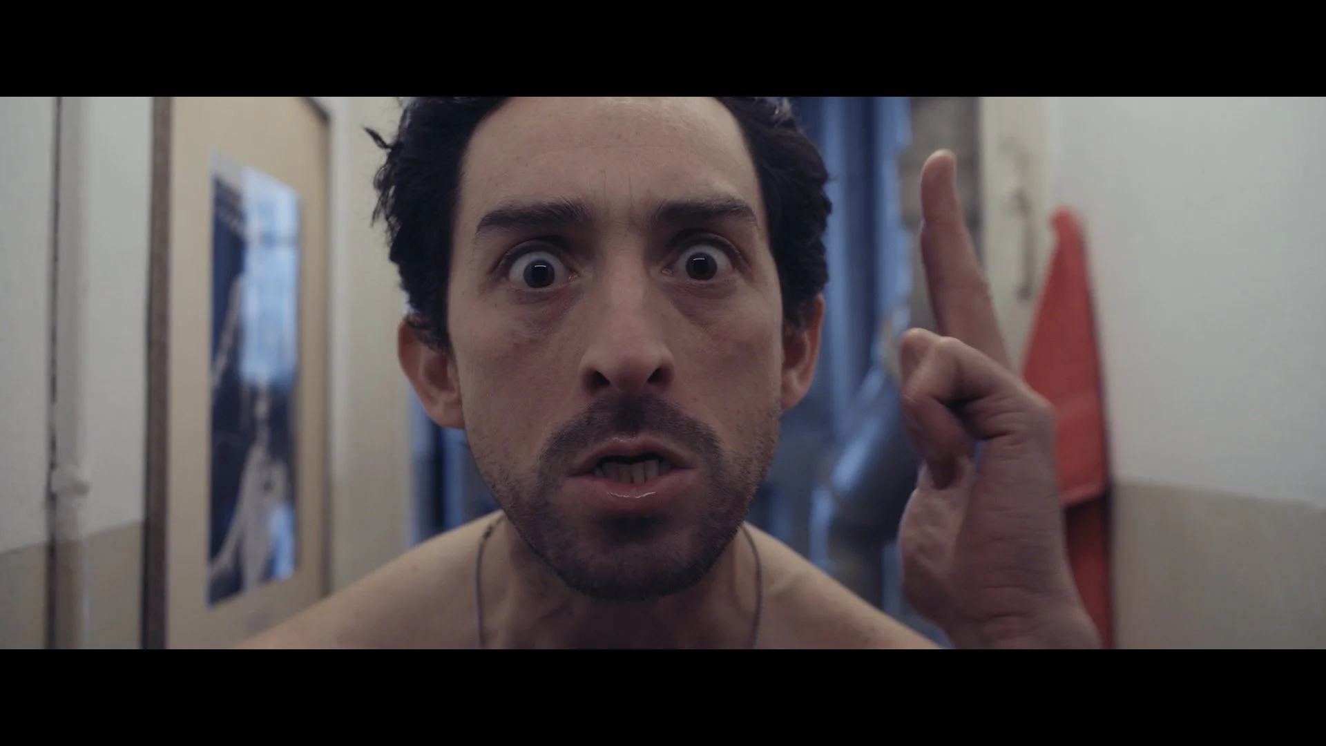 Nahaufnahme eines Mannes mit verwüstetem Gesicht und offenem Mund, der auf die Kamera zeigt, in einem Flur mit Fotos an den Wänden.