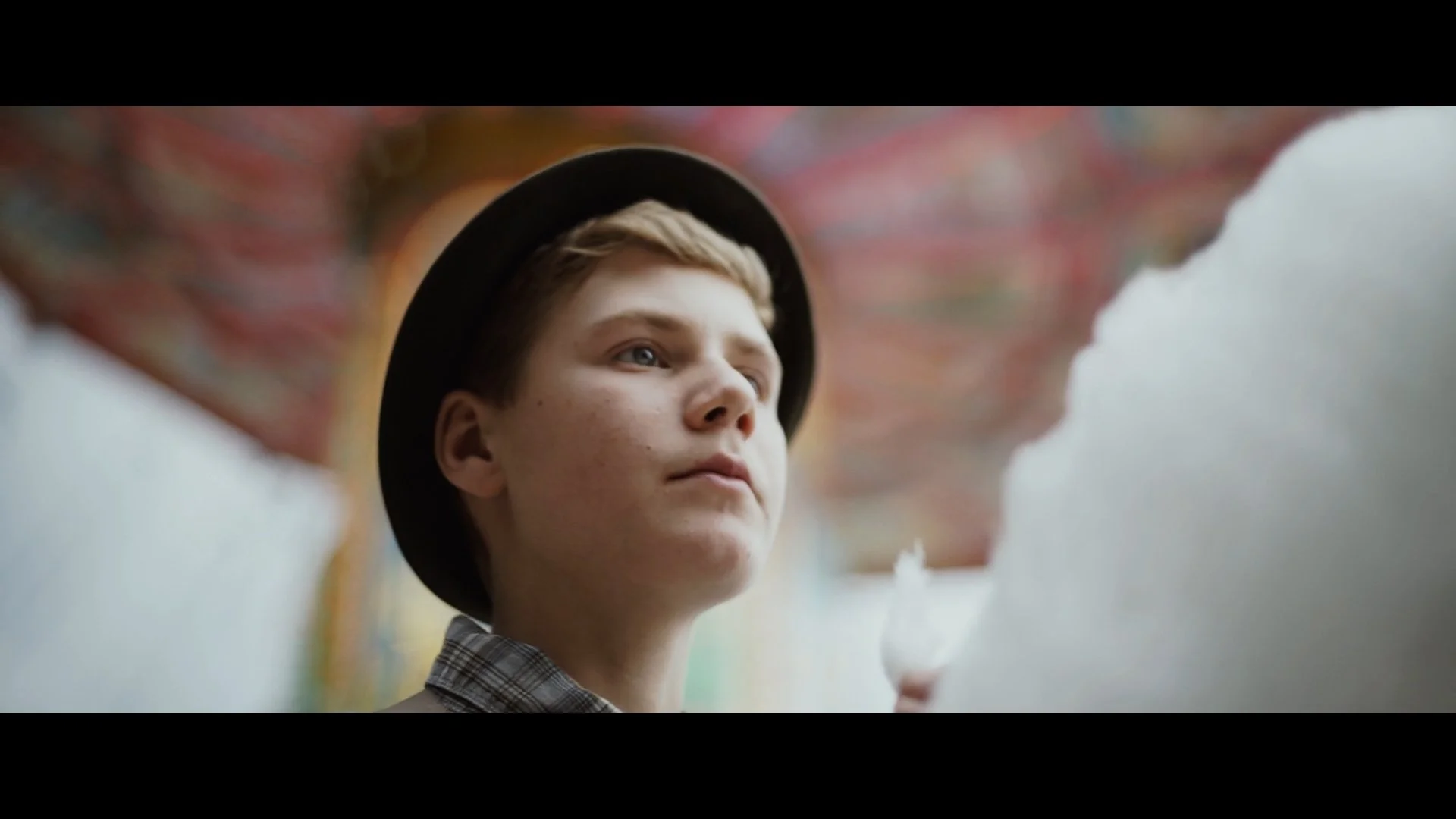 Ein junger Mann mit light Haart, trägt einen schwarzen Hut und schaut nach oben, im Hintergrund ein bunter, verwirrender Himmel, möglicherweise eine illusionäre Szene.