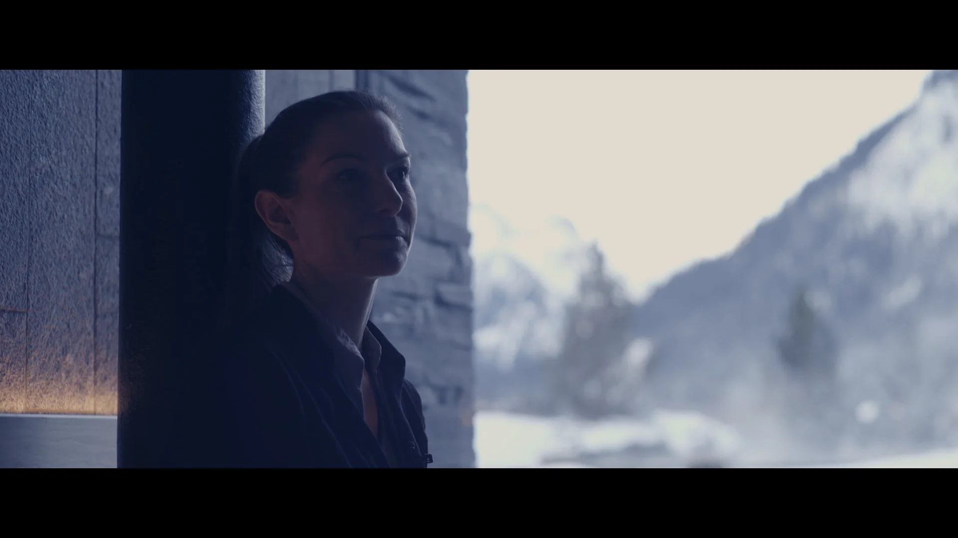 Frau sitzt in einem dunklen Raum mit Blick nach draußen, im Hintergrund ein verschneites Bergpanorama.