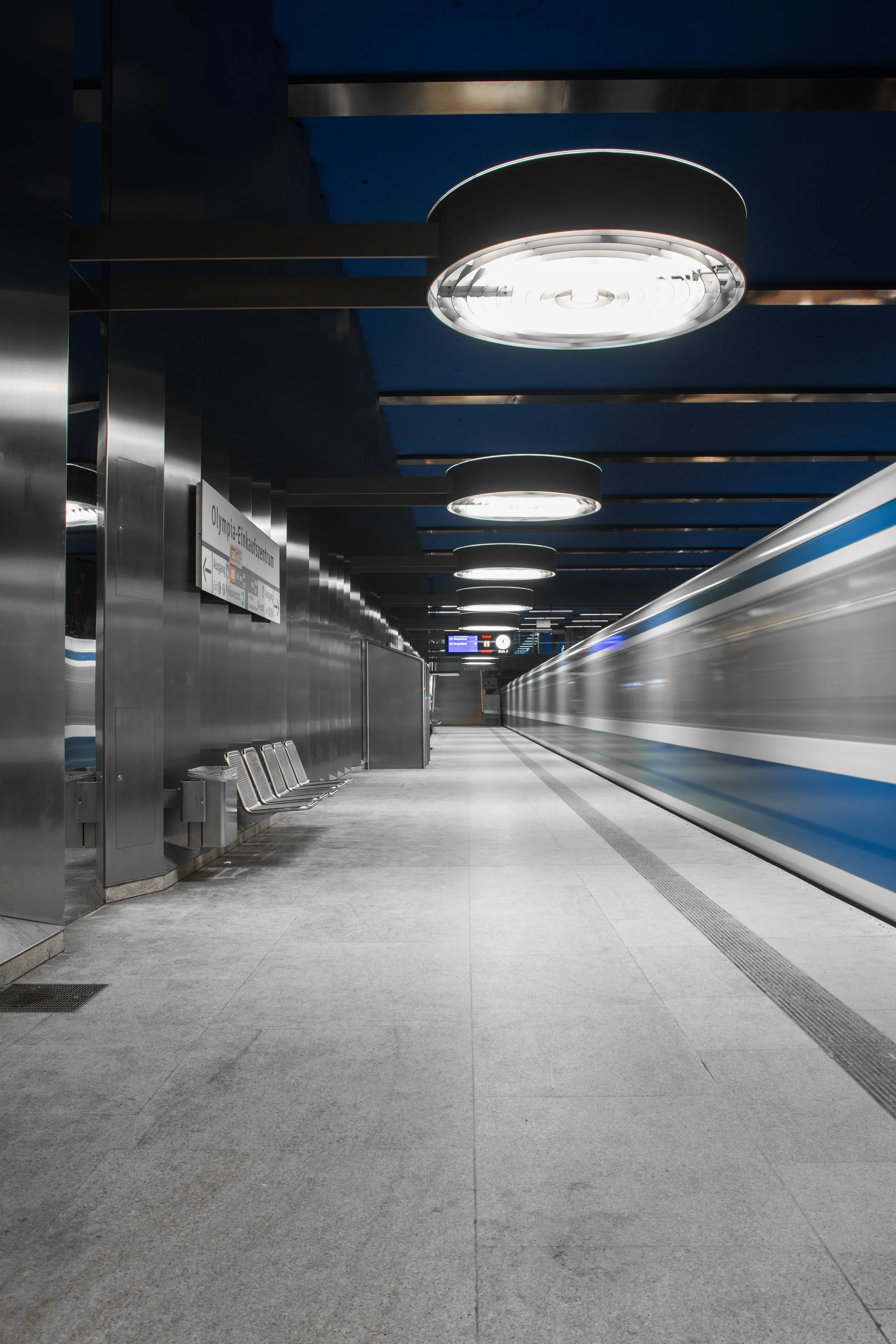 Leerer U-Bahnhof mit grauem Boden, modernen Sitzgelegenheiten, Wand- und Deckenverkleidungen aus Metall, beleuchteten runden Deckenleuchten und einem vorbeifahrenden Zug.