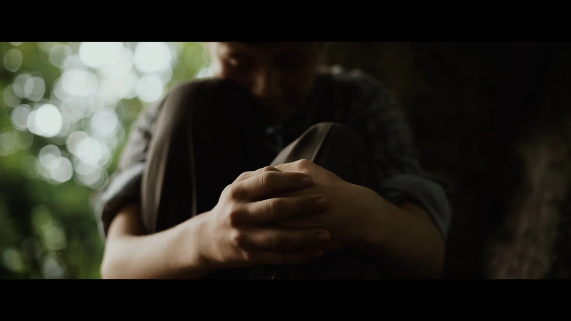 Nahaufnahme der Person, die ihre Knie umarmt und nachdenklich wirkt, mit unscharfem Hintergrund aus grünem Blätterdach.