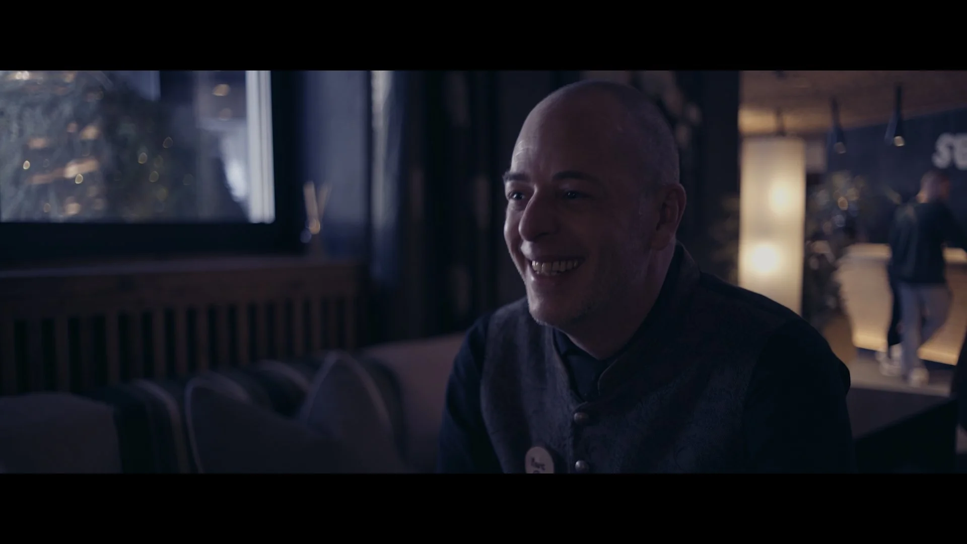 Ein lachender, glatzköpfiger Mann in einem dunklen Raum, vermutlich einem Restaurant oder Café, im Hintergrund sind weitere Personen und eine beleuchtete Bar.