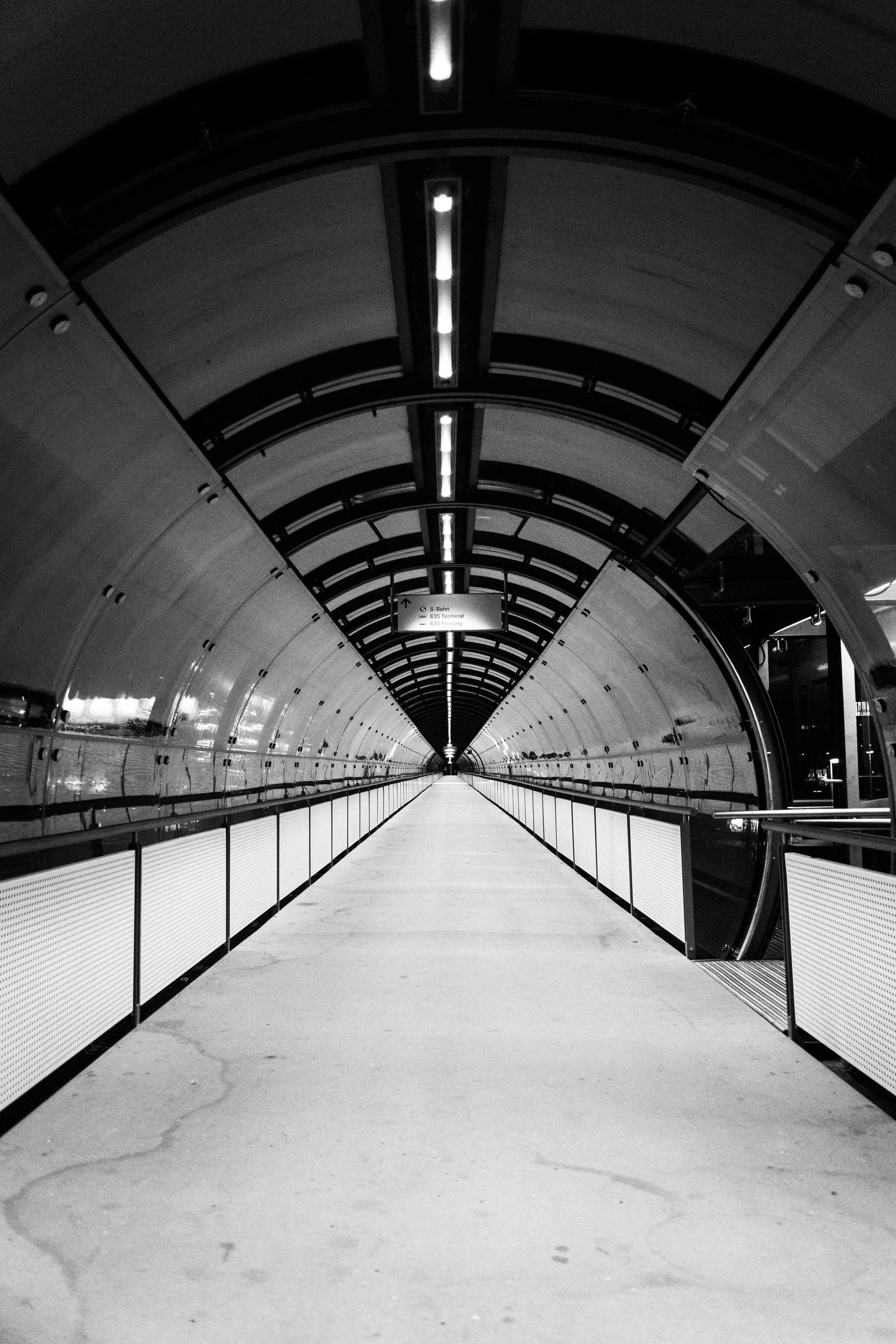 Ein schwarzer und weißer Tunnel mit Beleuchtung und Handläufen, wahrscheinlich eine Unterführung in einer U-Bahn-Station.