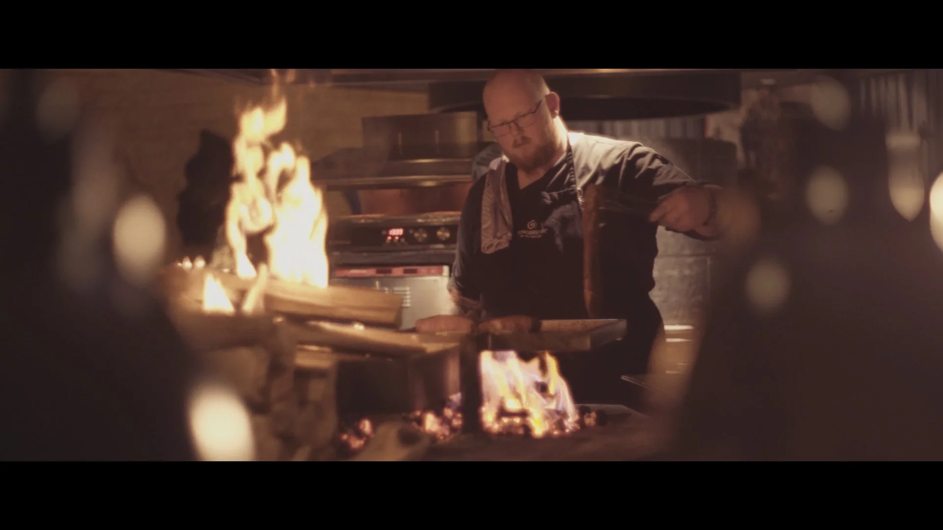 Koch in einer professionellen Küche, der mit einem Holzbrett hantiert, während Flammen aus dem Ofen schlagen.