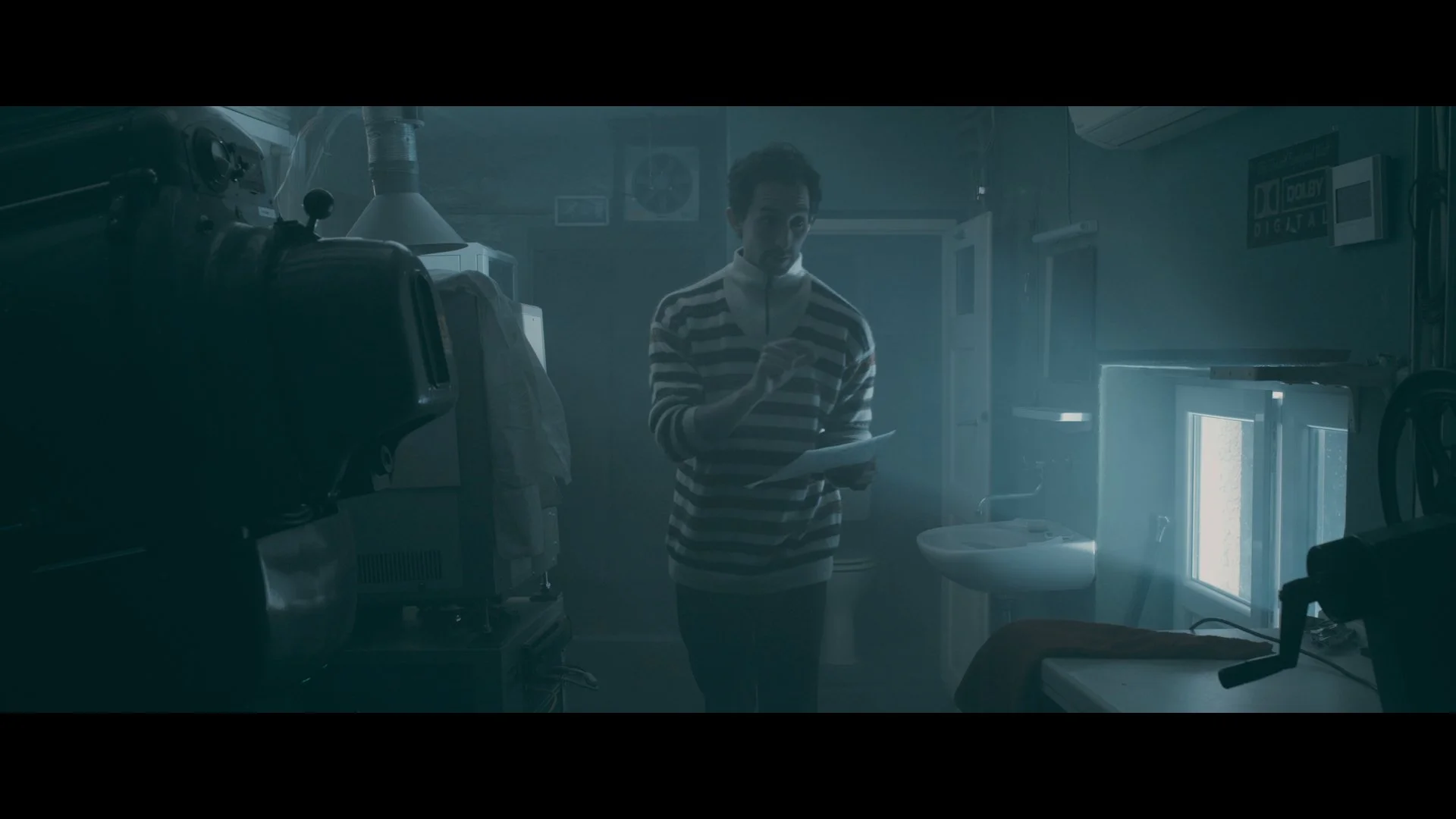 Ein Mann in einem gestreiften Pullover steht in einem dunklen Raum, liest ein Stück Papier und zeigt auf es.