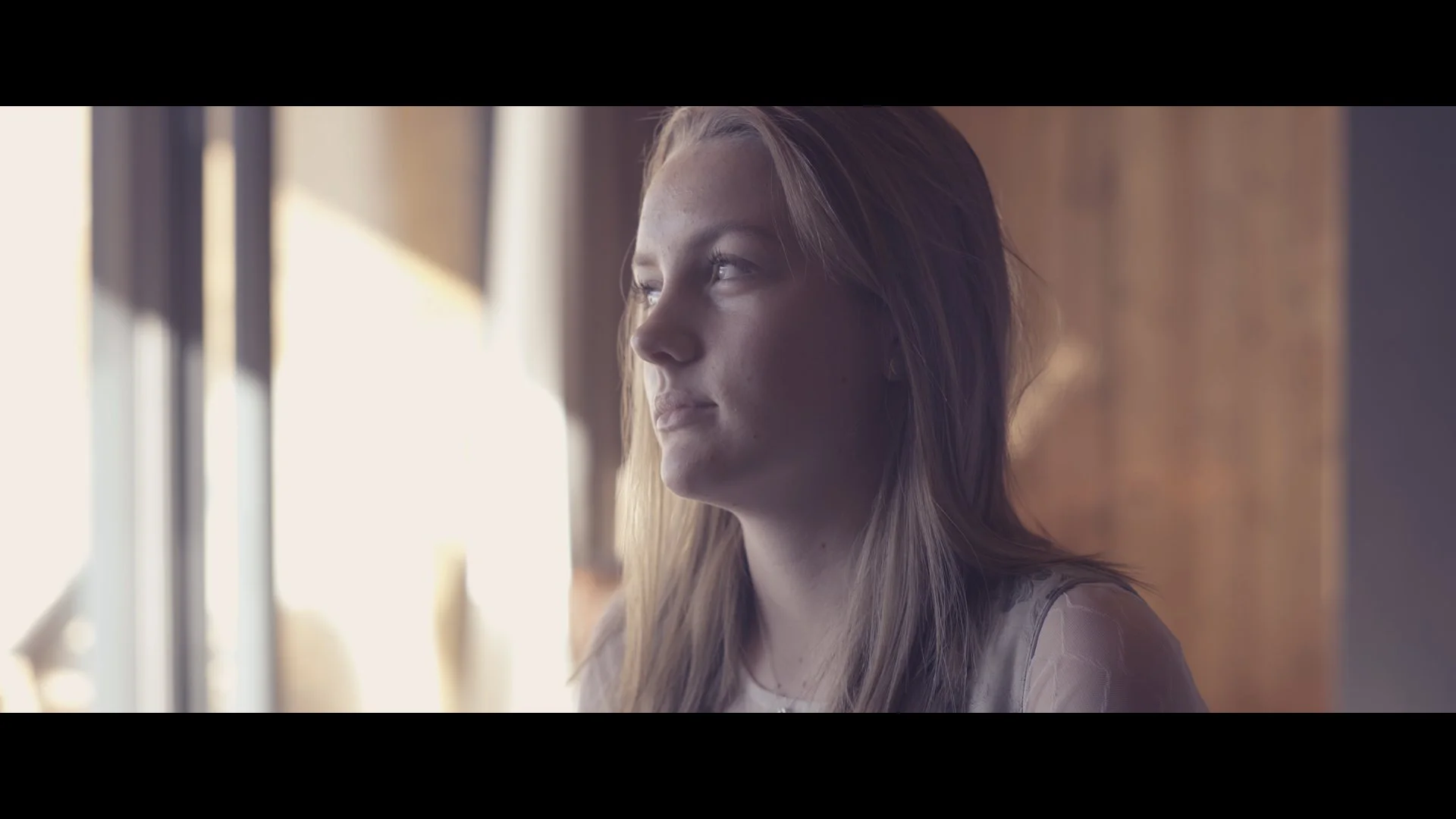 Nahaufnahmen eines jungen Mädchens mit langen, braunen Haaren, das nach draußen schaut, während Sonnenlicht durch ein Fenster fällt.