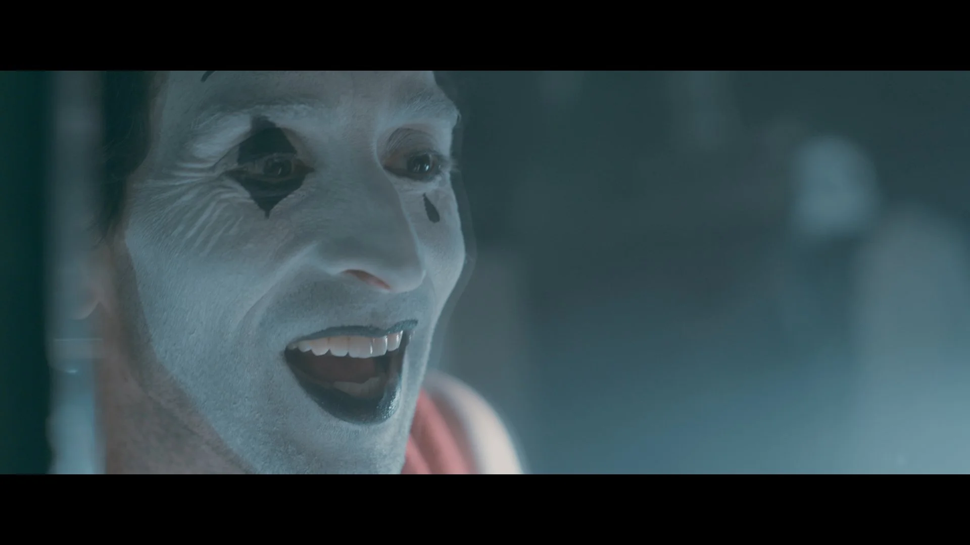Nahaufnahme eines Mannes mit clownartigem Make-up, lachend oder schreien, in einem dunklen, nebligen Umfeld.