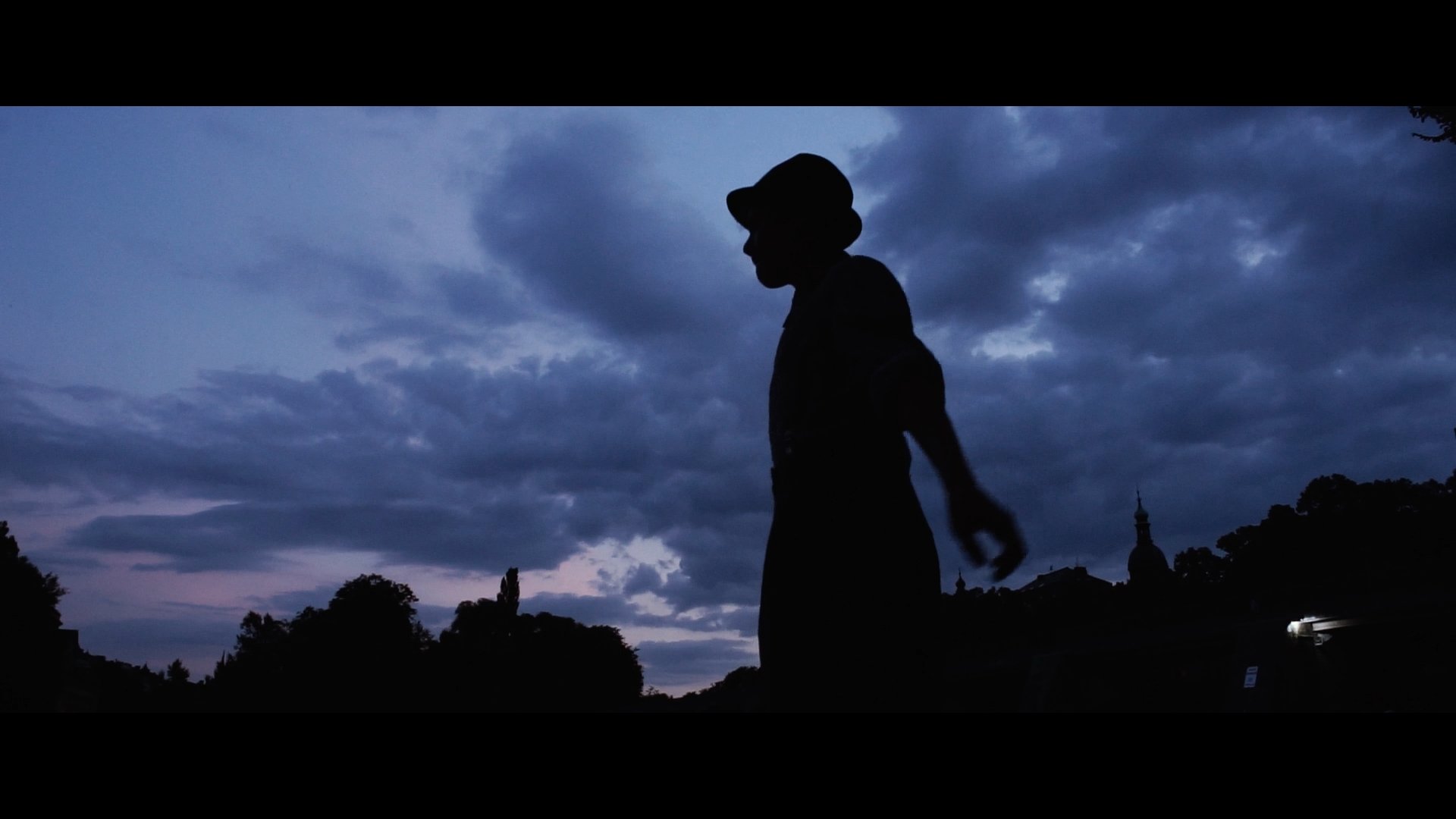 Schatten eines Mannes mit Hut im Abendhimmel mit Wolken, Bäumen und einem Turm im Hintergrund.