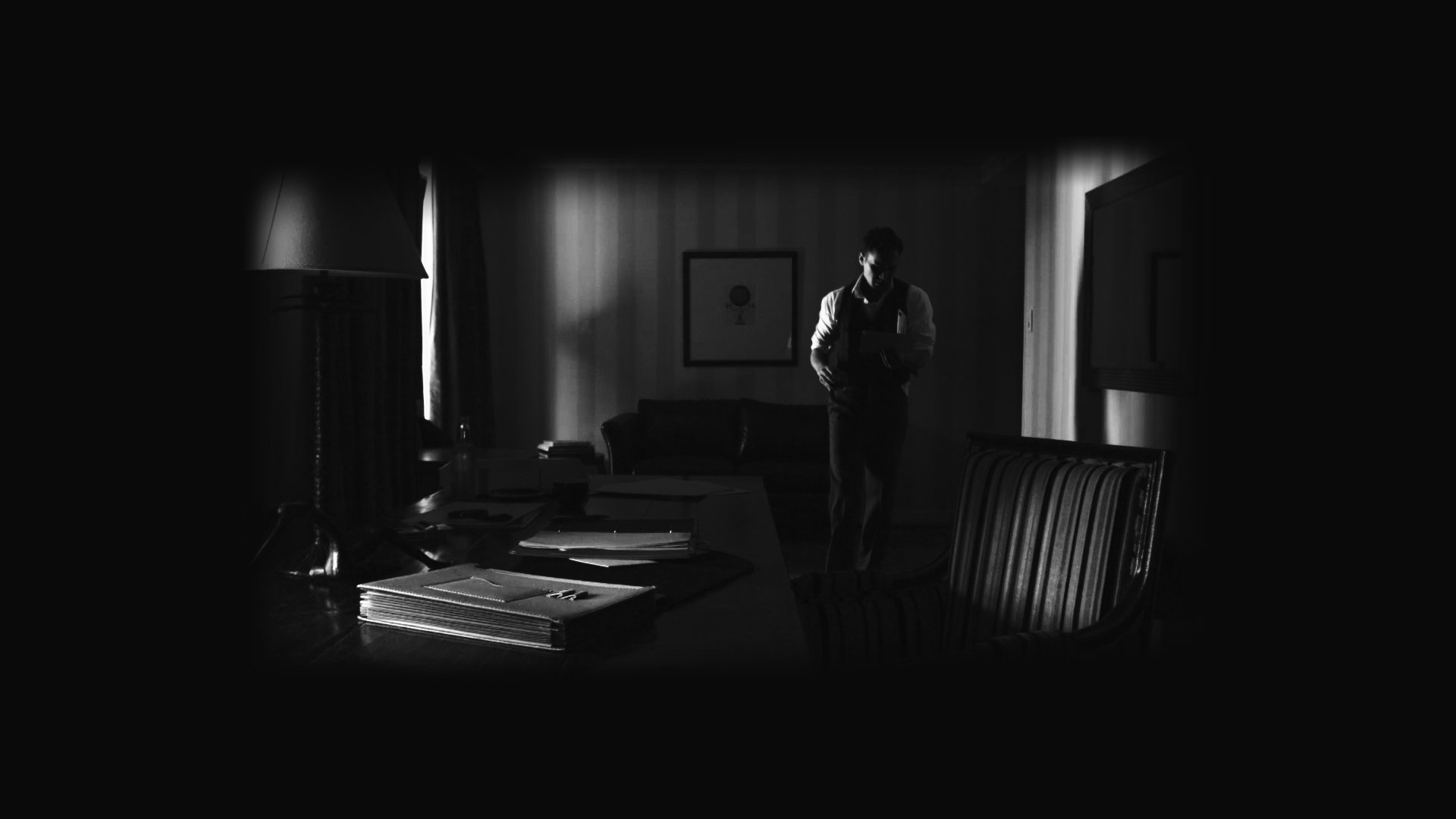 Ein Schwarz-Weiß-Bild zeigt eine Person, die in einem düsteren, schattenbedeckten Raum steht. Die Person hält etwas in den Händen und ist im Hintergrund zu sehen, während der Raum mit Möbeln und Büchern ausgestattet ist.