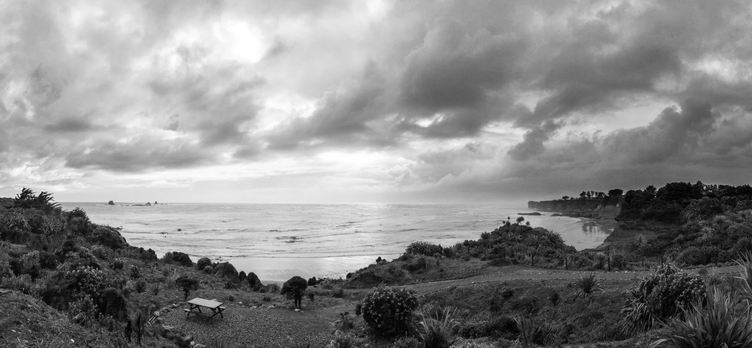 Panorama einer Küstenlandschaft mit Strand, Meer und bewölktem Himmel in Schwarz-Weiß. Es gibt Pflanzen und Bäume auf den Hügeln im Vordergrund, eine Bank und einen kleinen Weg. Klippen und Felsen im Wasser im Hintergrund.