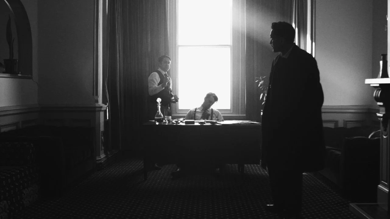 Drei Männer in einem formellen Raum mit einem großen Fenster im Hintergrund, zwei sitzen an einem Schreibtisch, einer steht. Es ist ein Schwarz-Weiß-Bild im Vintage-Stil.