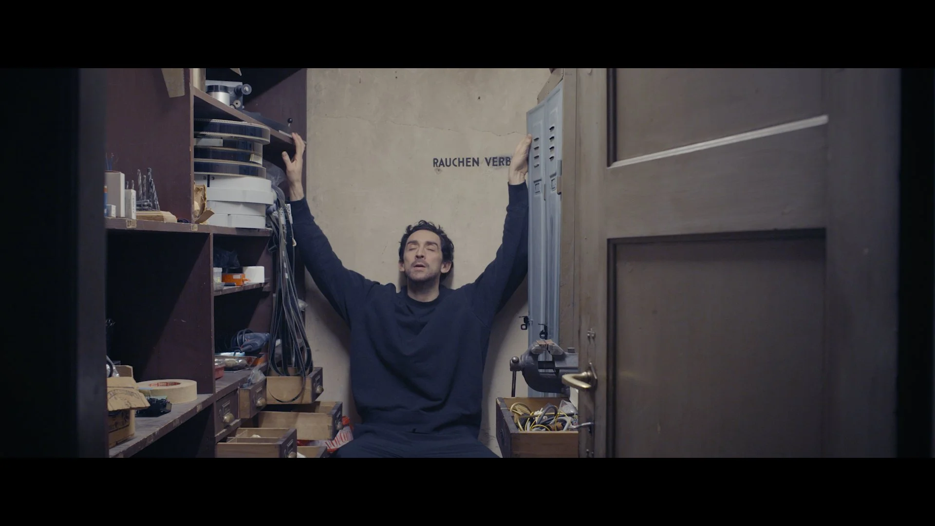 Ein Mann sitzt im Schrankraum mit ausgestreckten Armen, die Augen geschlossen, umgeben von Werkzeugen und Bauteilen, hinter ihm eine Wand mit dem Schriftzug 'RAUCHEN VERBOTEN'.