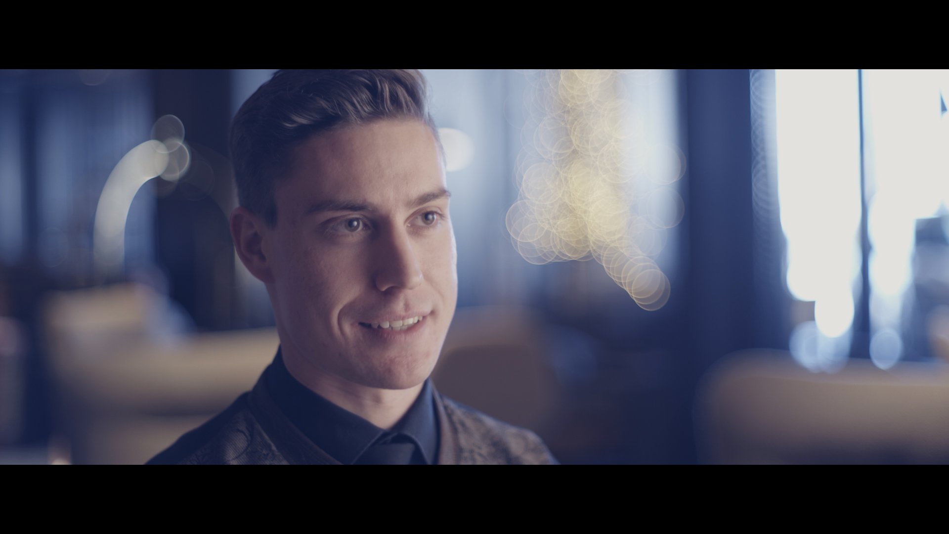 Junge Mann mit dunklem Hemd, lächelnd, im Innenraum mit bokeh-Hintergrund, Weihnachtslichter, moderne Einrichtung.