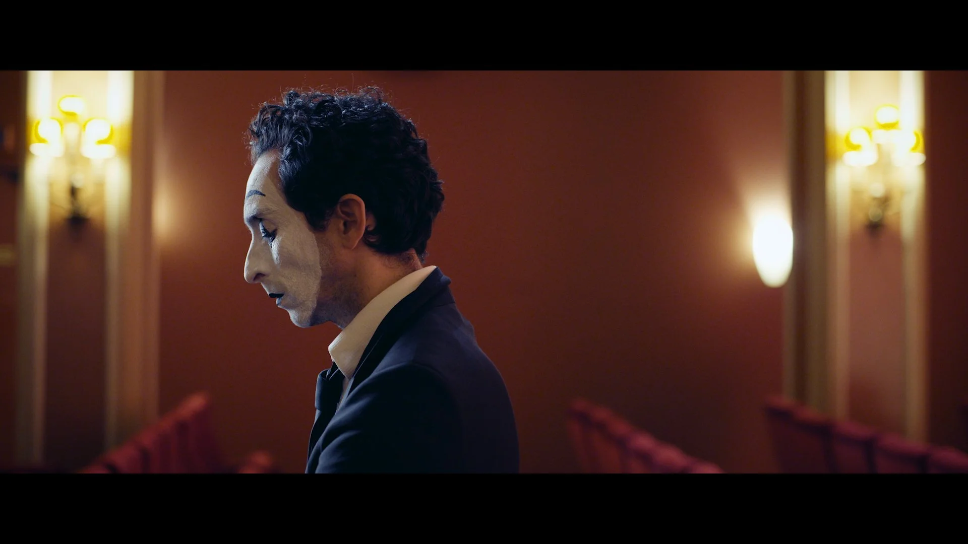 Ein Mann in einem schwarzen Anzug und weißem Hemd sitzt mit gesenktem Kopf in einem Theater oder Konzertsaal, der Raum ist gedämpft beleuchtet mit roten Wänden und warmem Licht.