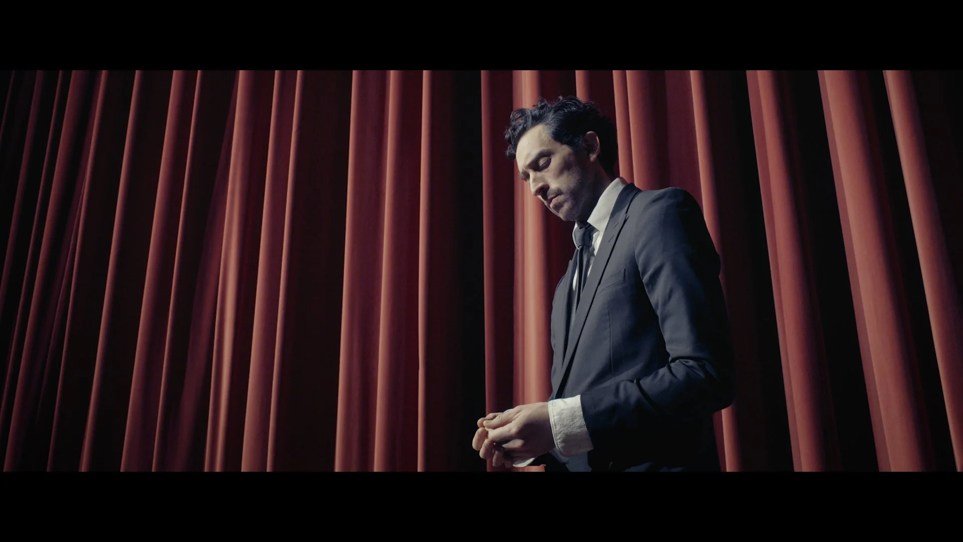 Ein Mann in dunklem Anzug steht vor roten Vorhängen, blickt nach unten und hält seine Hände gefaltet.