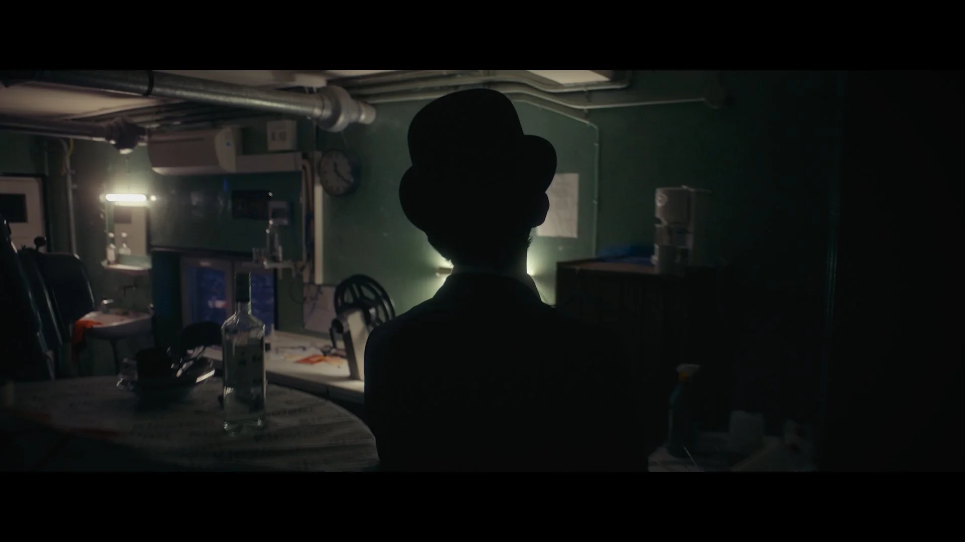 Schatten einer Person mit Zylinder im dunklen Raum, Blick auf einen Tisch mit Wasserflasche und diversen Gegenständen, im Hintergrund technische Geräte und grüne Wände.