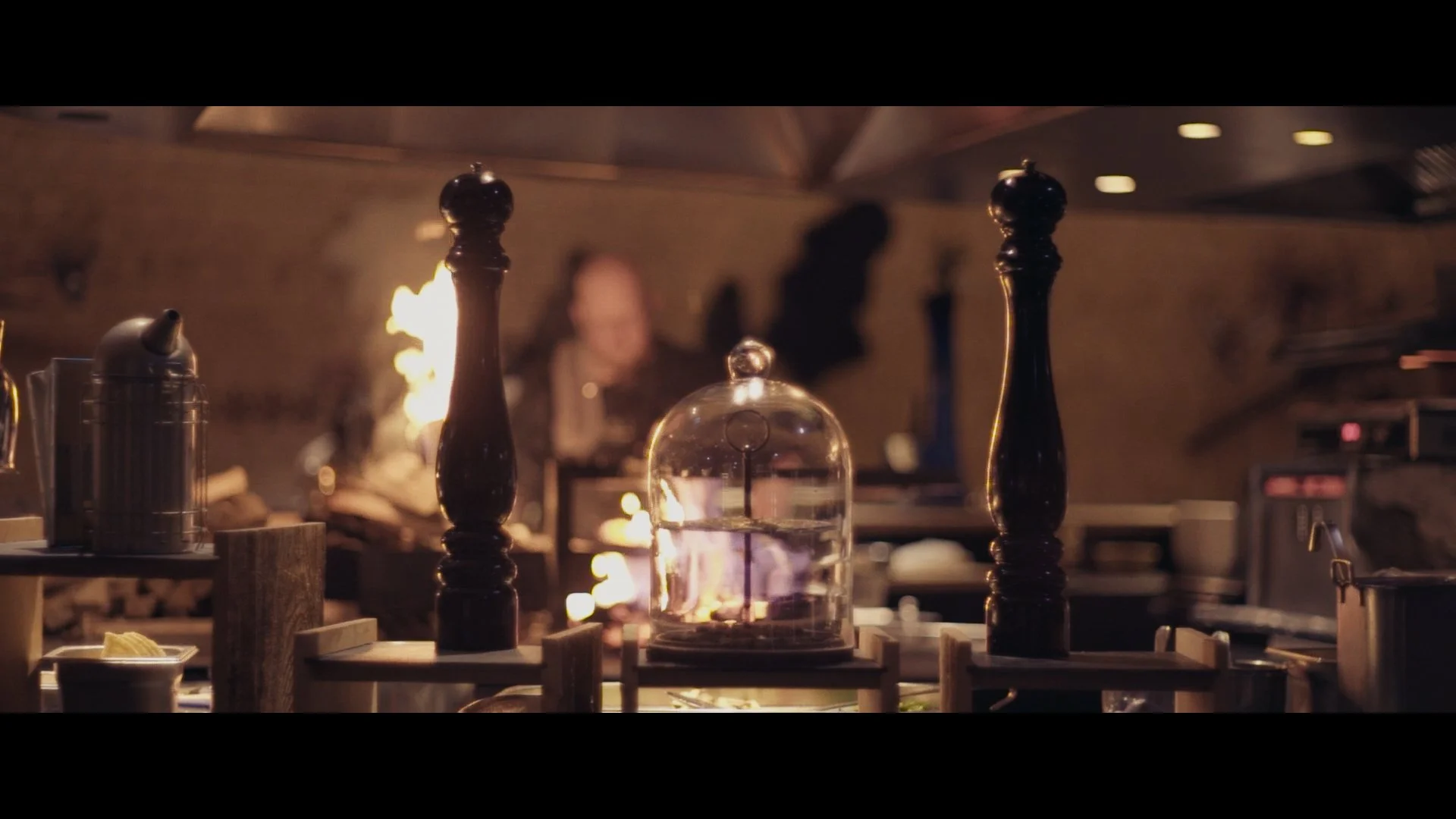 Blick auf eine Küchenarbeitsplatte mit Salz- und Pfefferstreuern, eine Glasglocke mit Kerzenlicht und einem Feuer im Hintergrund, die Szene wirkt warm und rustikal.