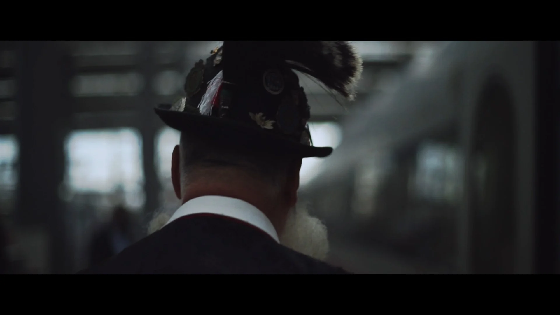 Rückansicht eines Mannes in traditioneller bayerischer Tracht mit Hut, dekoriert mit Feder und Abzeichen, in einem Hotel oder Bahnhofshalle, gedämpftes Licht