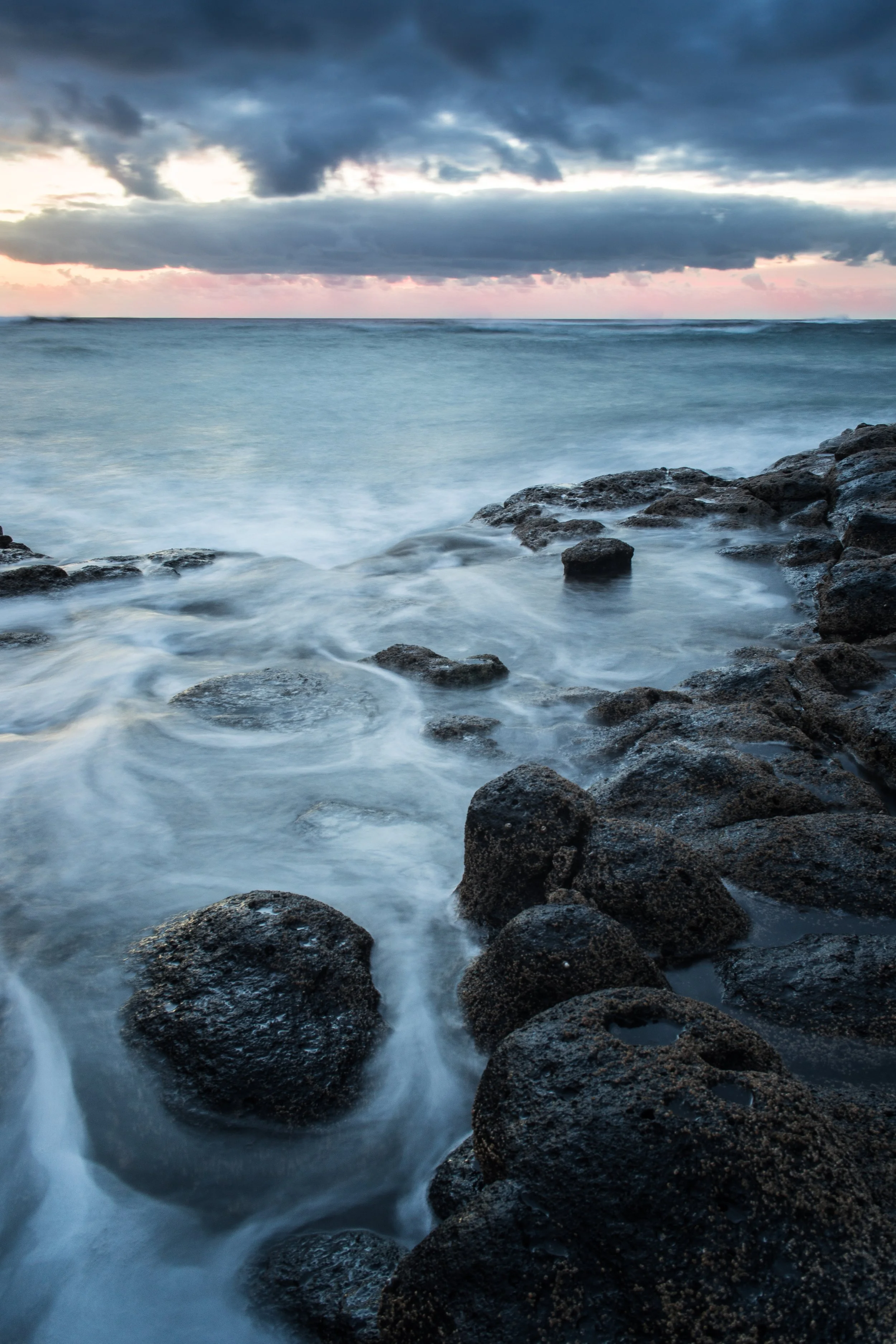 Meer mit dunklen Steinen im Vordergrund, stürmischer Himmel und Sonnenuntergang im Hintergrund.