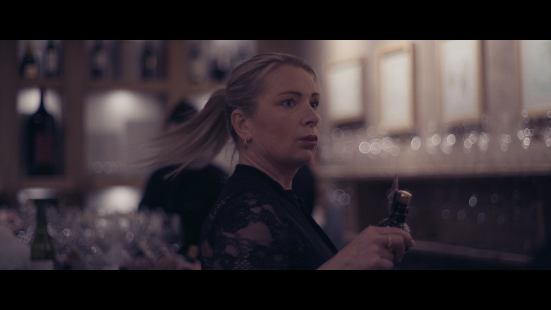 Eine Frau mit blondem Haar in einem dunklen Kleid hält eine Flasche in der Hand, im Hintergrund sind Gläser und Flaschen in einer Bar sichtbar.