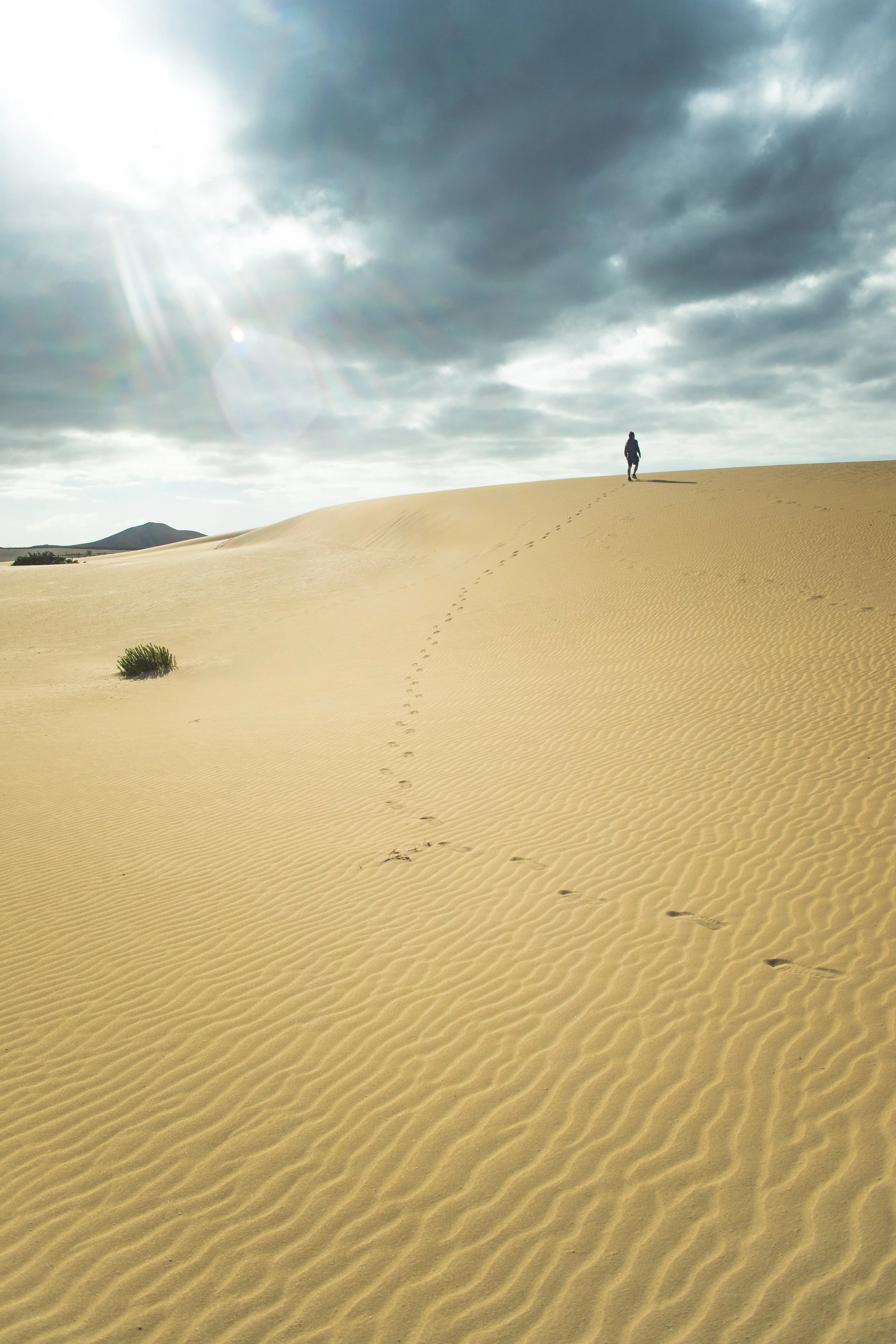 Ein Mensch läuft auf einer sandigen Düne in einer Wüste, vom Himmel strahlen Sonnenstrahlen durch Wolken, mit entfernten Hügeln im Hintergrund.