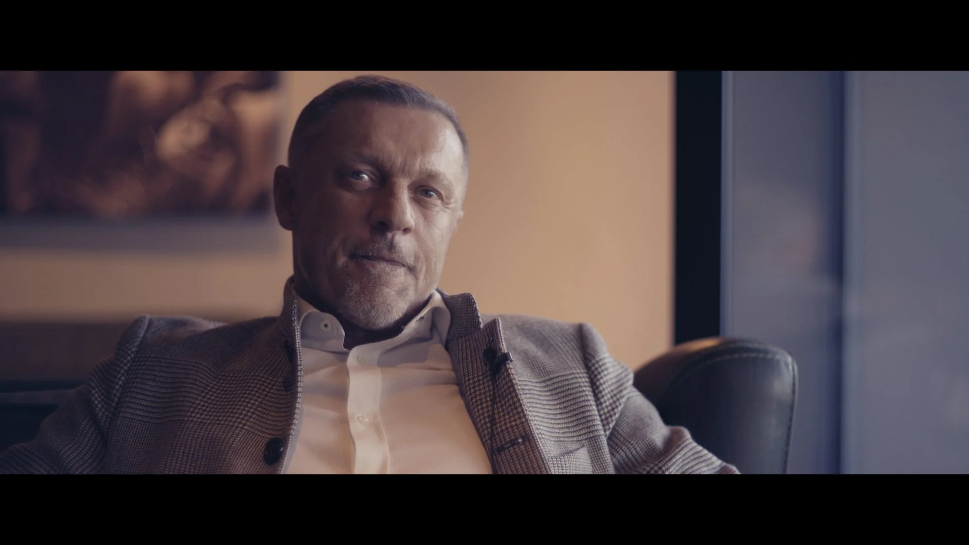 Ein Mann sitzt in einem Sessel, trägt einen karierten Blazer und einen weißen Hemd, schaut nach vorne, im Hintergrund eine Wand mit einem Kunstwerk.