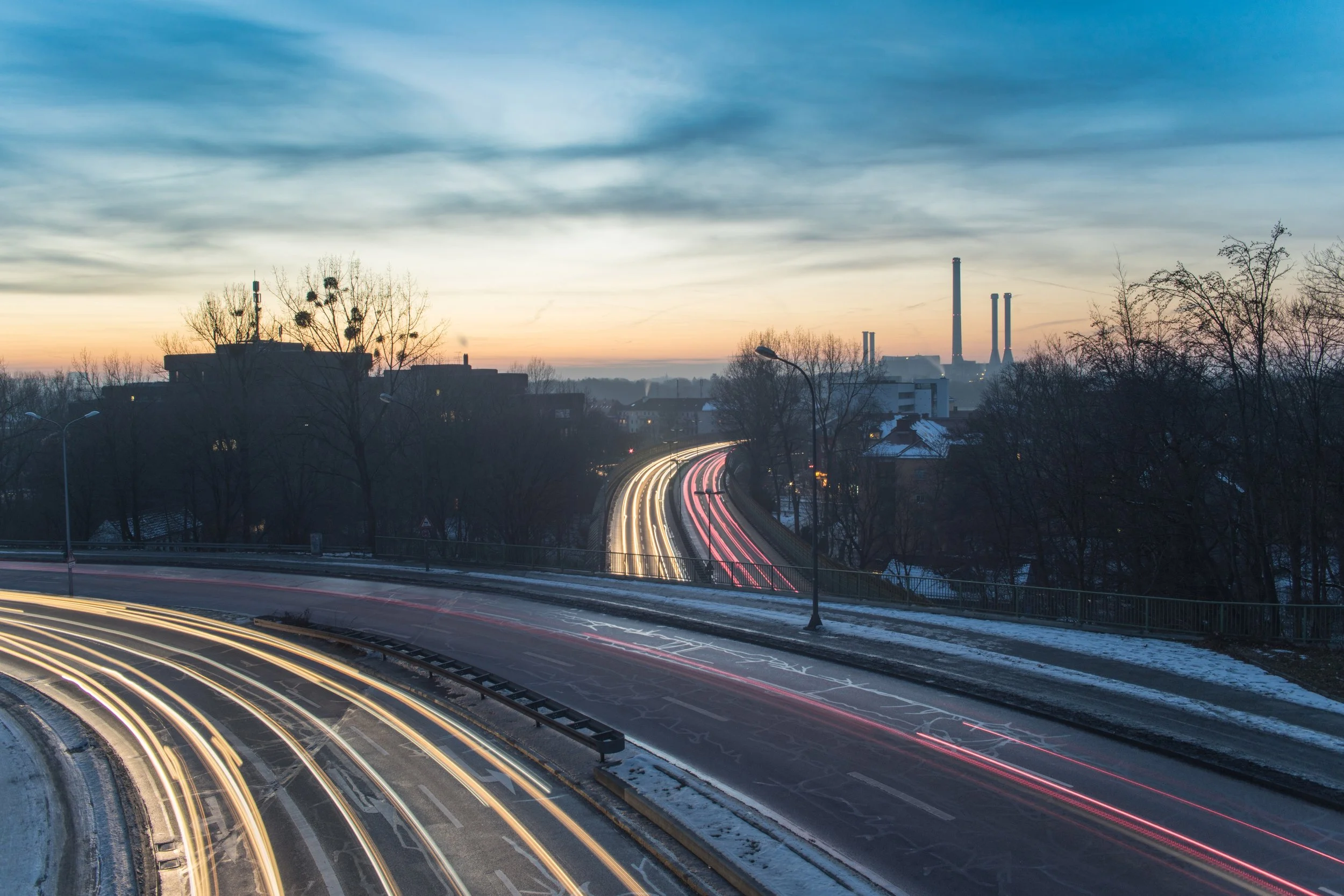 Abendlicher Blick auf eine Schnellstraße mit Lichtspuren, umgeben von Bäumen und Gebäuden, im Hintergrund eine Stadt mit Fabrikschornsteinen und Wolken im Himmel