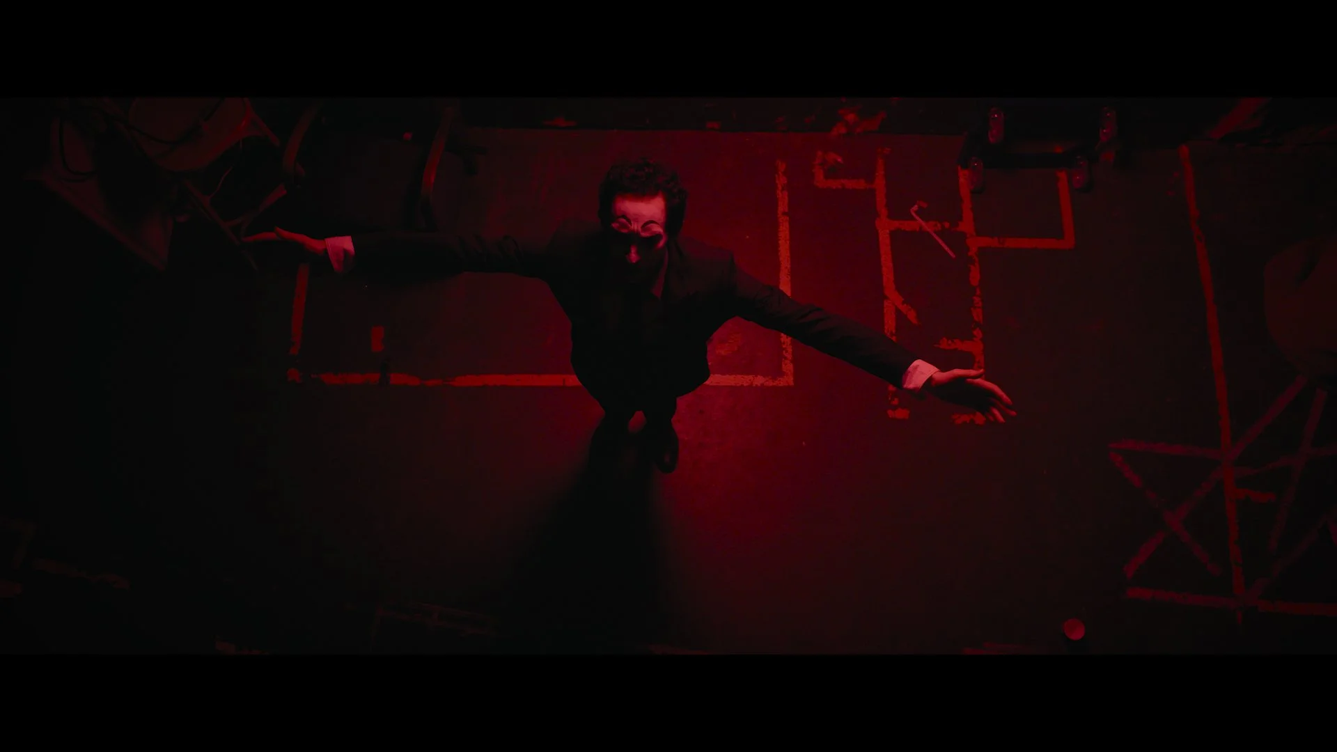 Persönlichkeit in schwarzem Anzug, maskiert, steht in einem dunklen Raum mit rotem Licht, umgeben von Stühlen, mit Aufsicht von oben aufgenommen.