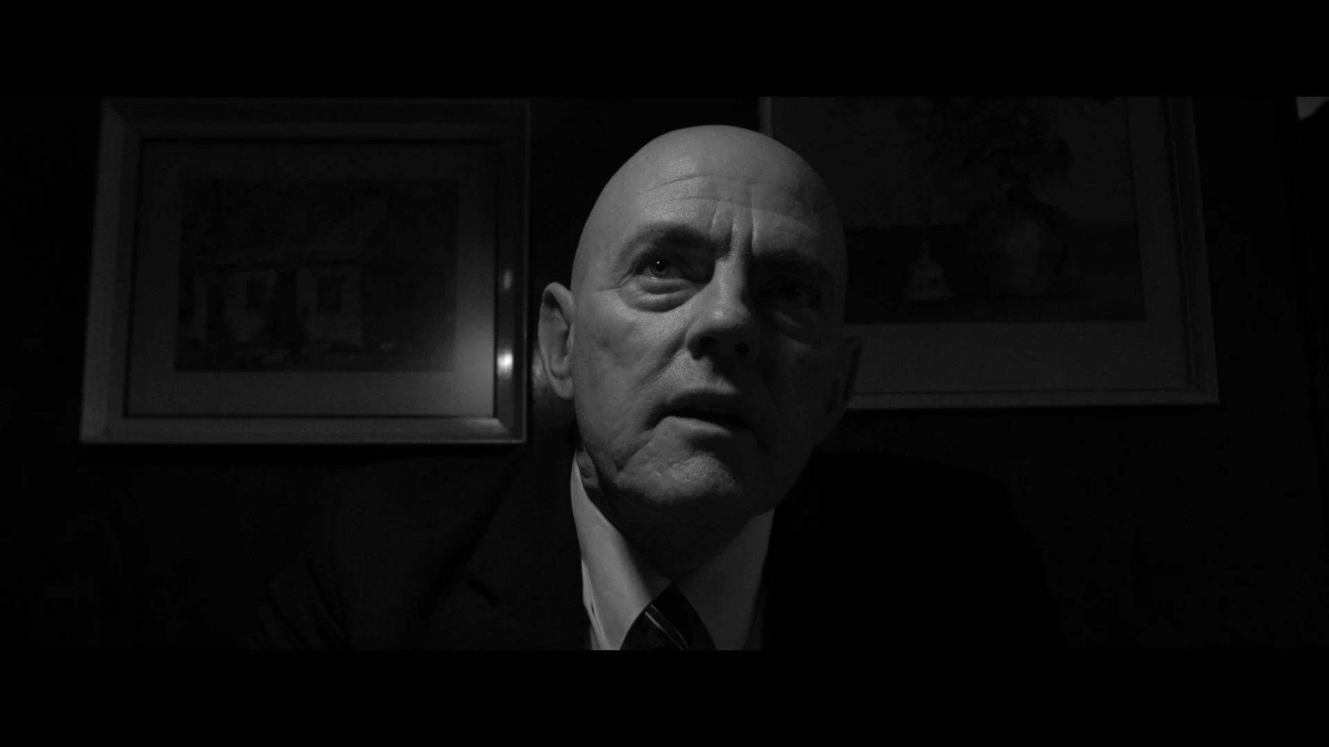 Schwarz-Weiß-Bild eines Mannes in einem Anzug, der nach oben schaut, mit fokussiertem Blick, im Hintergrund zwei Gemälde an der Wand.