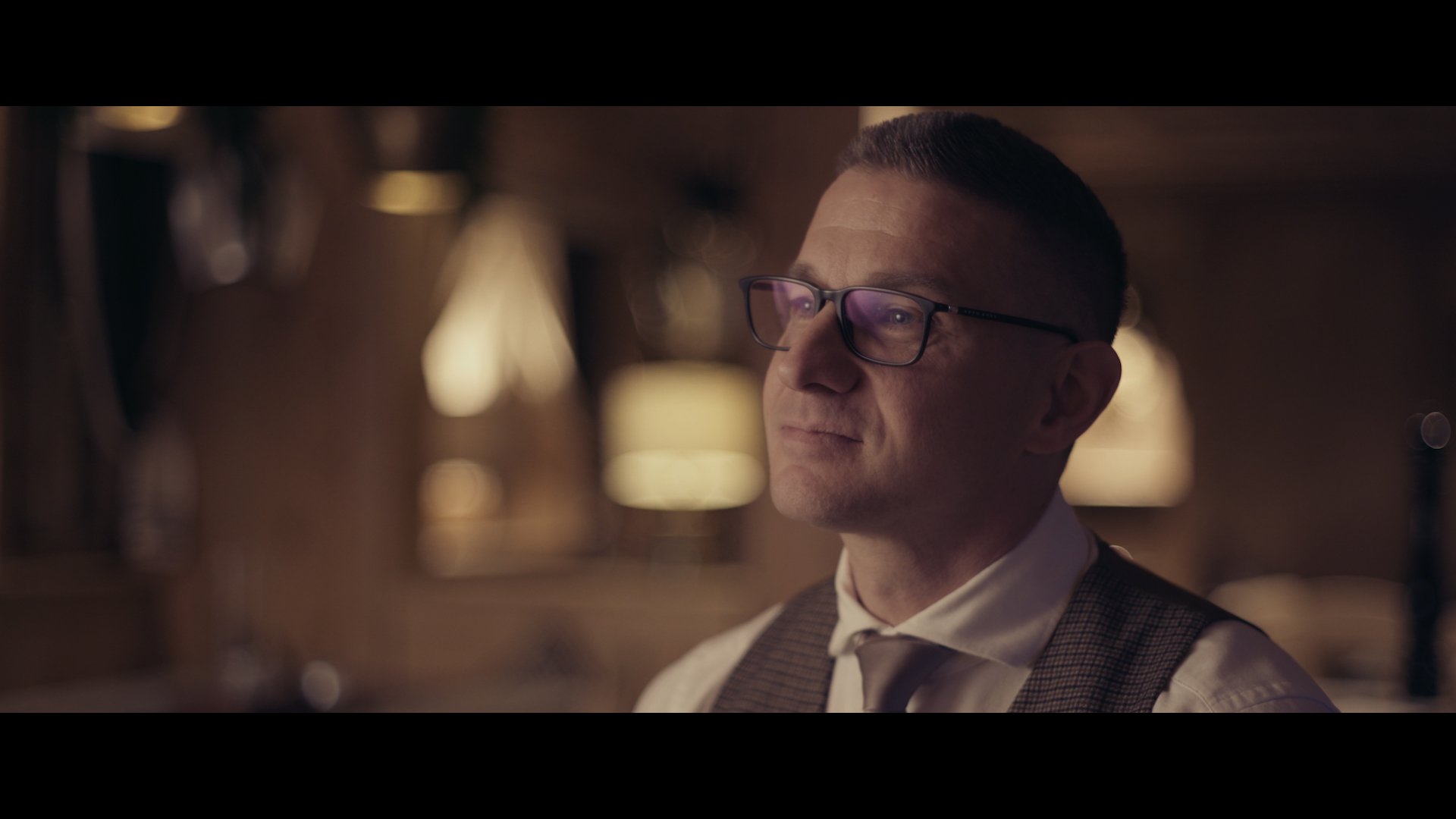 Nahaufnahme eines Mannes mit Brille, der einen nachdenklichen Blick hat, in einer warm beleuchteten Umgebung.