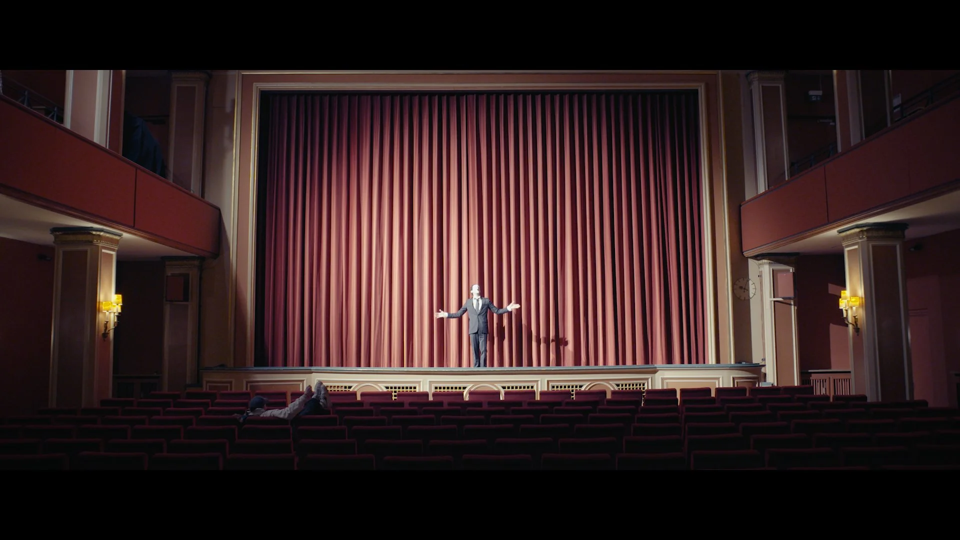 Ein Mann steht auf einer Theaterbühne in einem großen Saal mit roten Vorhängen, er trägt einen schwarzen Anzug und gestikuliert mit offen Armen, während einige wenige Zuschauer im Saal sitzen.