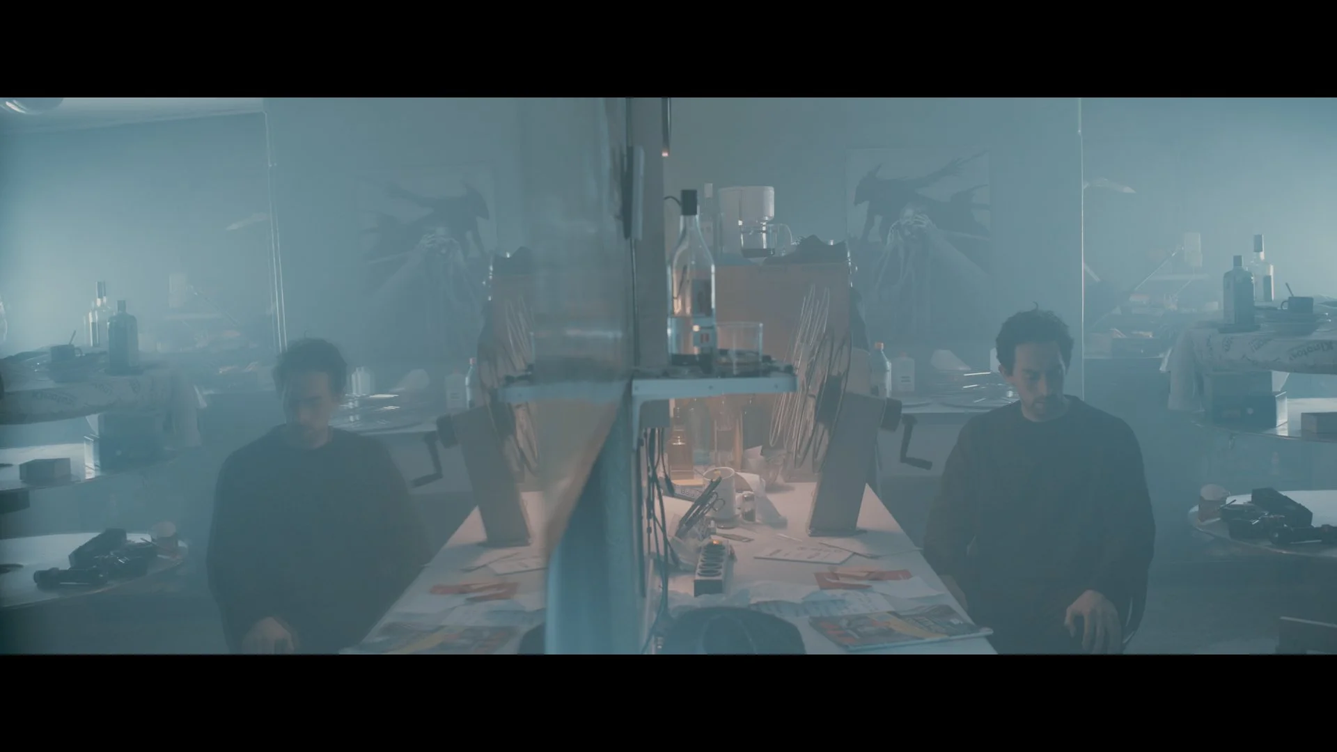 Zwei Männer sitzen in einem verrauchten Raum, umgeben von Tischen mit Büchern, Papieren, Flaschen und Geräten, hinter einer vernebelten Glasscheibe.