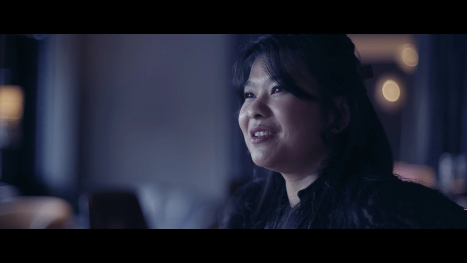 Nahaufnahme einer lachenden asiatischen Frau mit langen schwarzen Haaren, im Hintergrund unscharfe Lichter, wahrscheinlich in einem Innenraum.