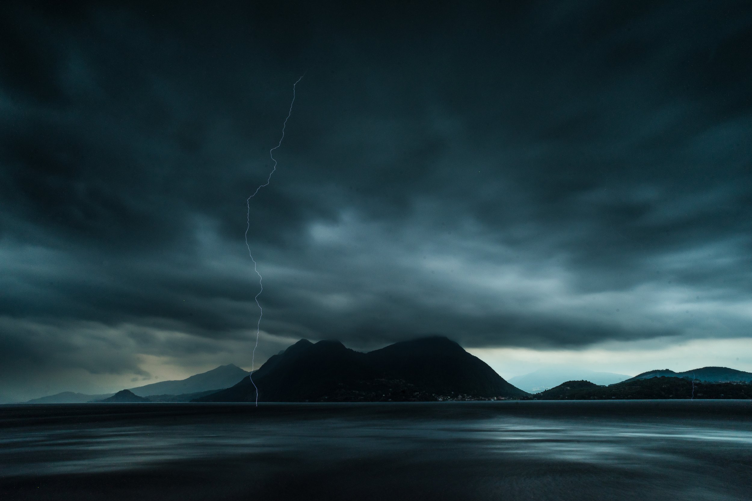 Sturm über einem See mit dunklen Wolken, einem Blitz und Bergen im Hintergrund.