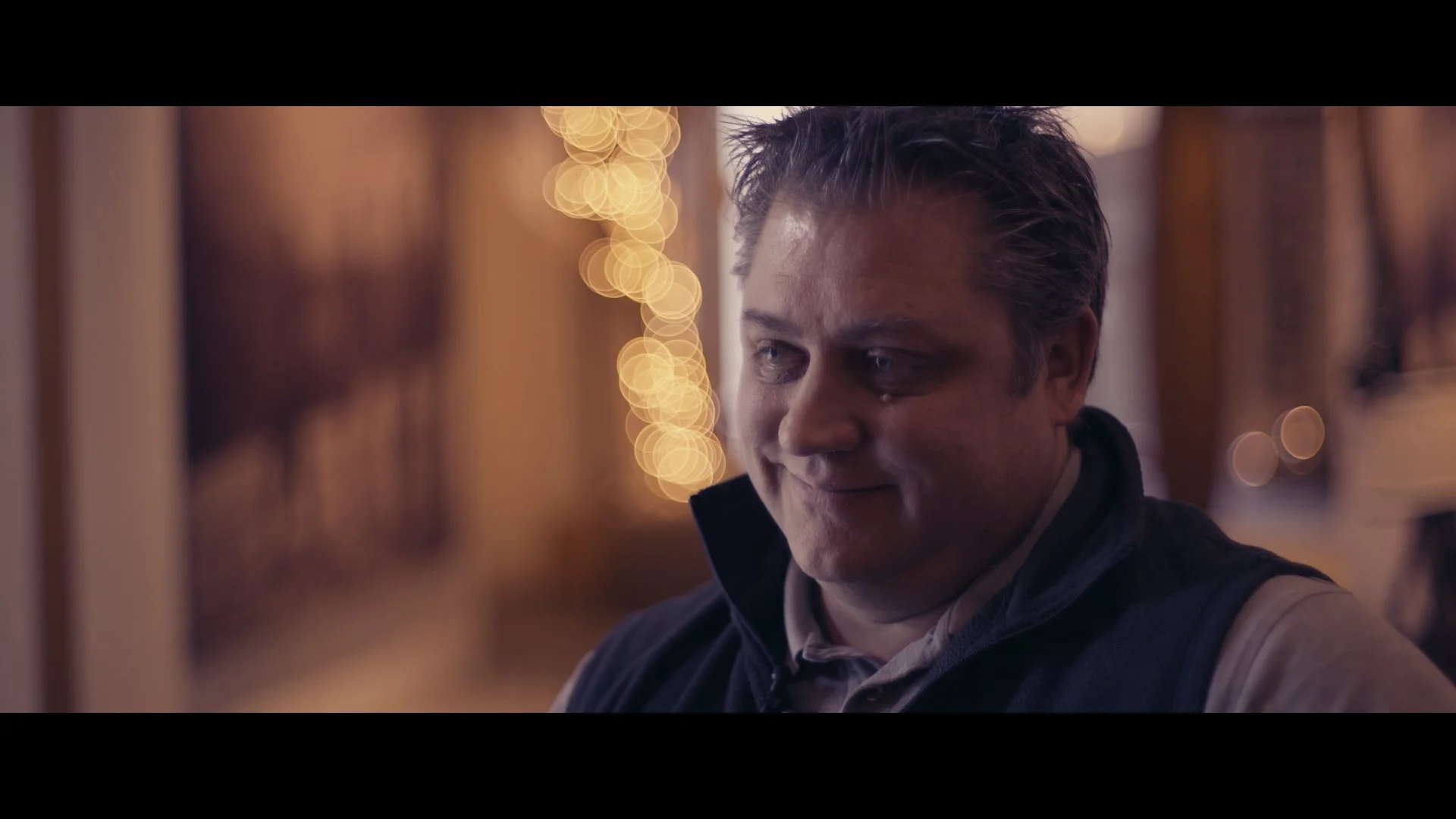 Ein Mann mit braunen Haaren, trägt ein dunkles Jackett und ein Hemd, steht in einem Raum mit bunter Beleuchtung im Hintergrund.