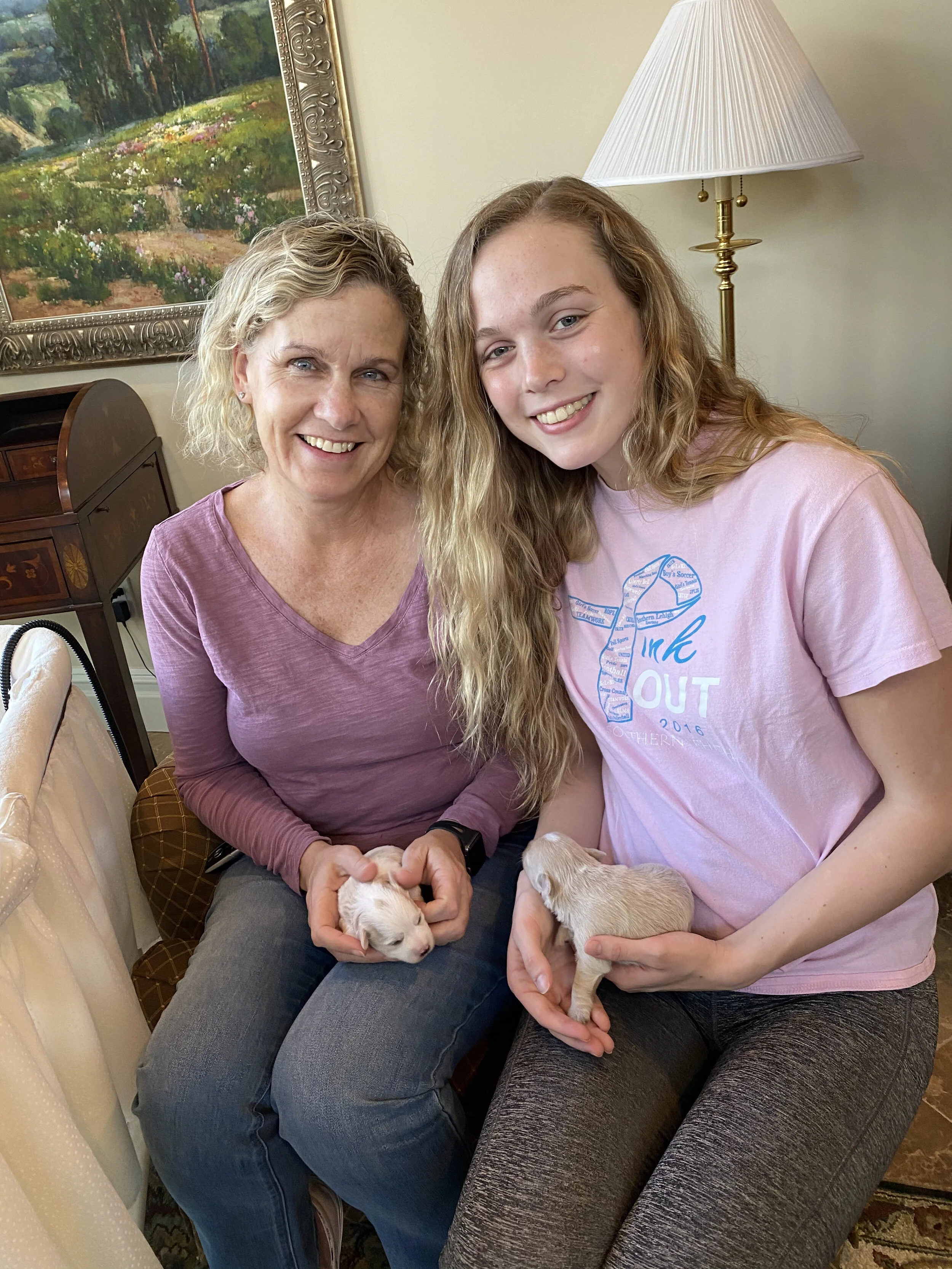 Two women, one older and one younger, smiling and holding puppies, sitting on sofas in a cozy room with a painting and a floor lamp in the background.