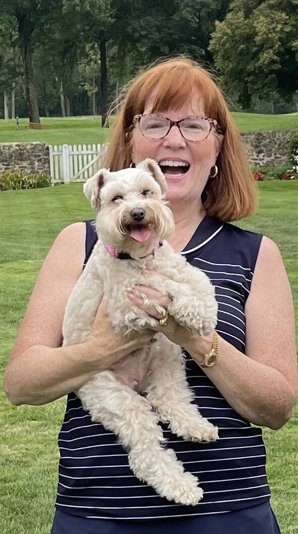 A woman with red hair and glasses smiling and holding a small, fluffy white dog outdoors on a green lawn with trees and a stone wall in the background.