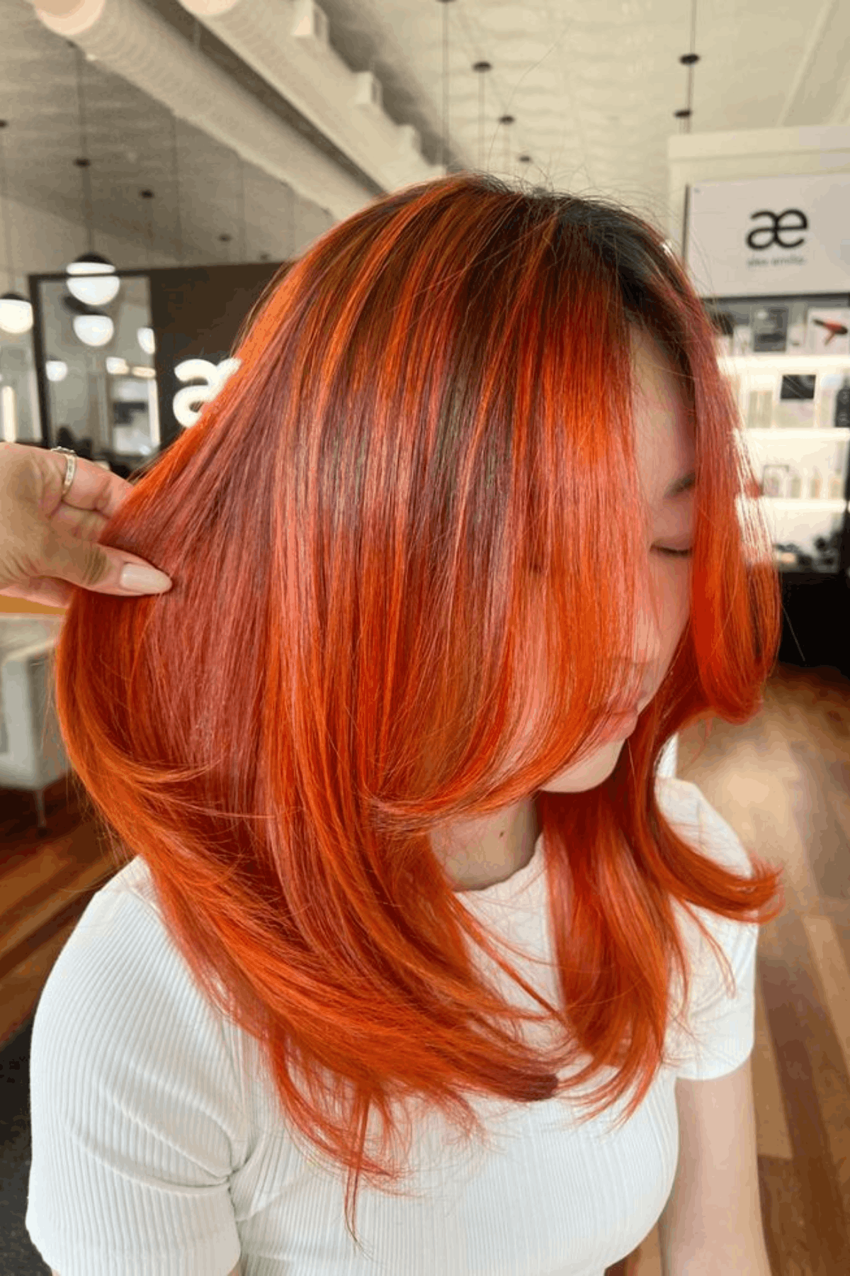Woman with layered medium-length hair in vivid sunset orange tones, blending bright orange with deeper copper for a bold, summery look.