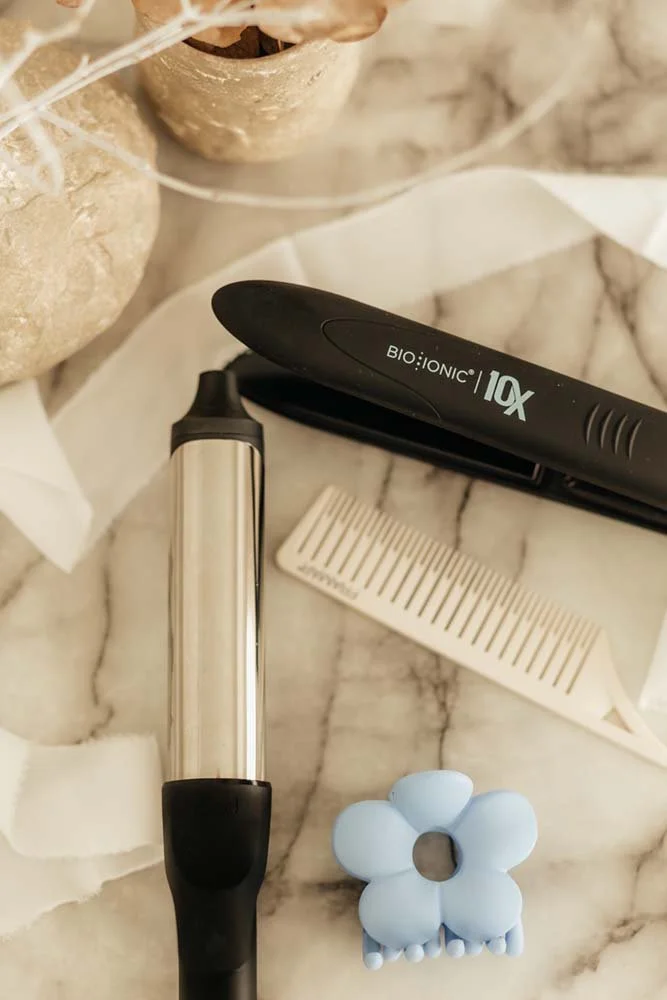 On a marble surface, Haiirology by Jen showcases an array of hair tools: a flat iron, a curling iron, a white comb, and a blue flower-shaped hair clip.