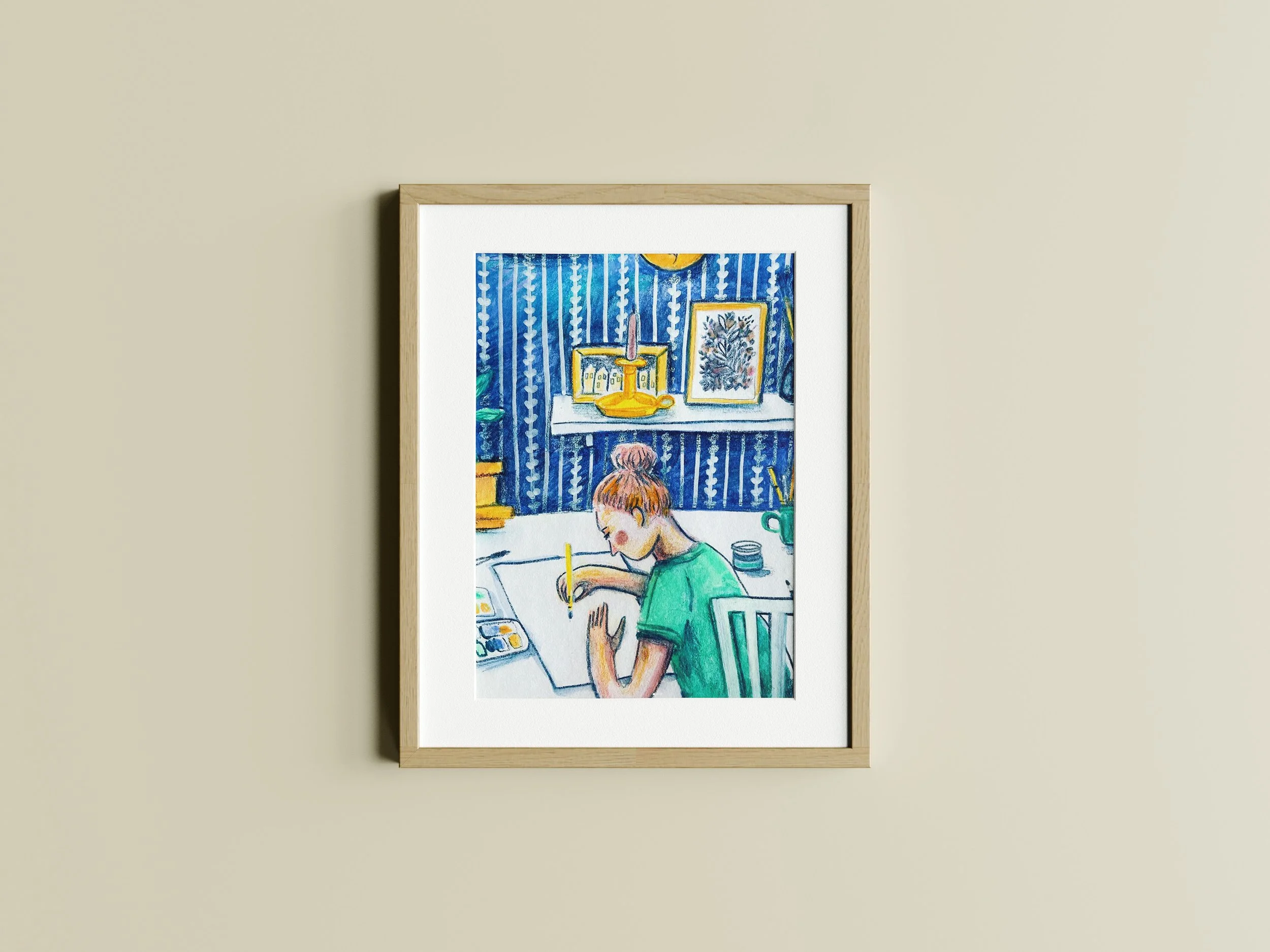 Framed artwork depicting a person drawing at a desk with blue patterned wallpaper, books, and plant, the artwork is made by illustrator Majken Enequist