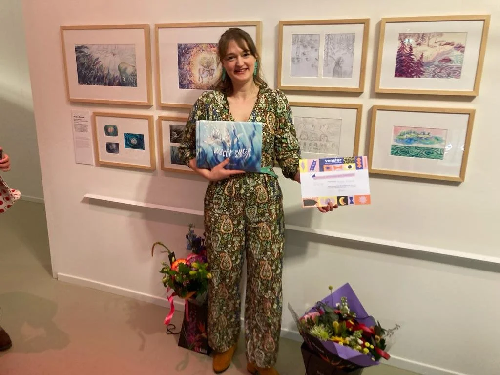 Woman standing in front of an exhibition wall displaying framed artworks. She is wearing a patterned outfit and holding a book and a certificate. Two bouquets of flowers rest on the floor next to her, the artwork is made by illustrator Majken Enequis