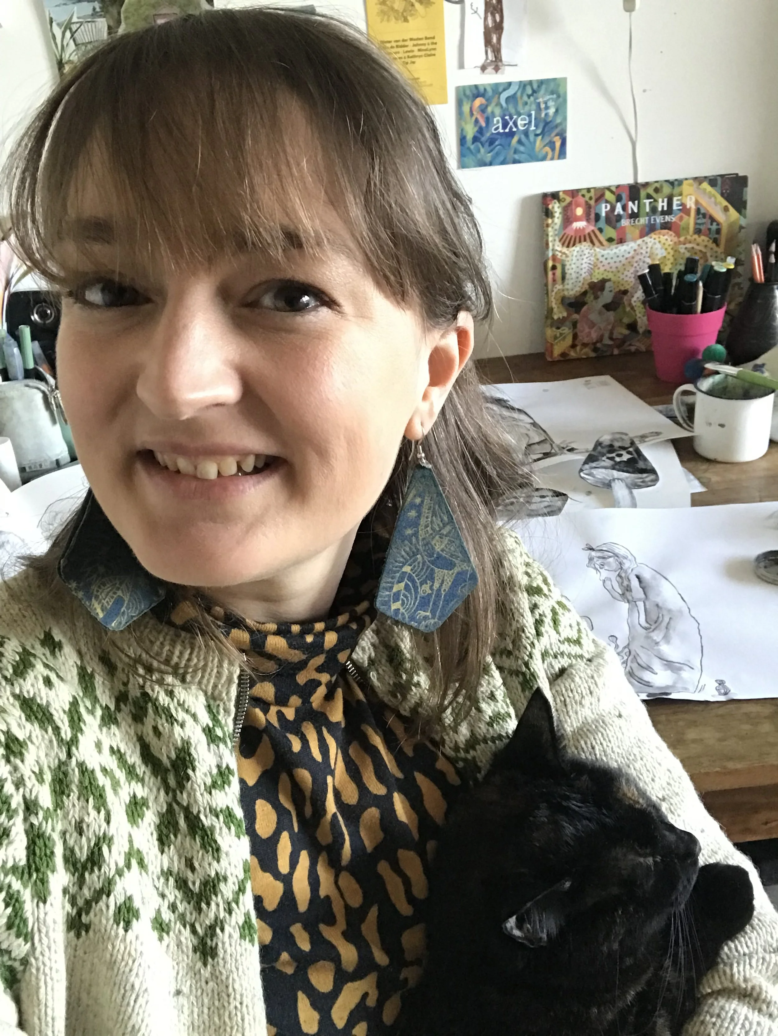 Person smiling while holding a black cat, wearing a patterned sweater and large earrings, in a room with art supplies and drawings on a desk, the artwork is made by illustrator Majken Enequist
