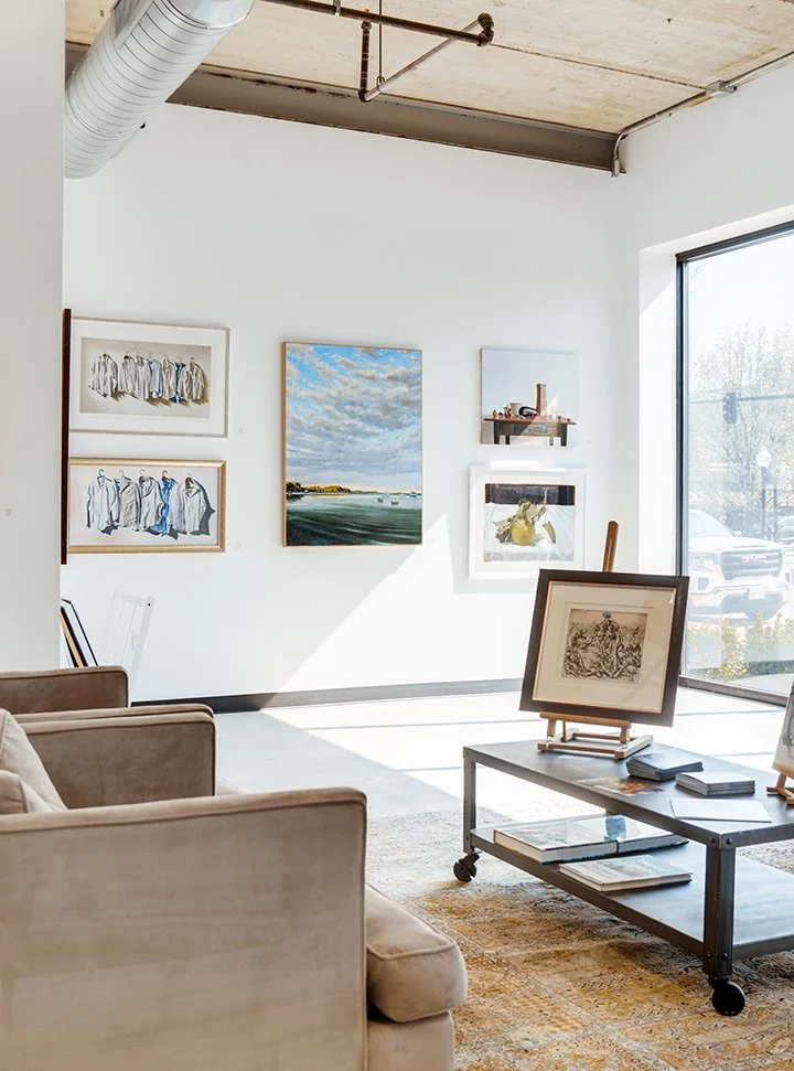 An art gallery or studio with paintings on the white walls, a beige sofa, a coffee table with more artwork, a large window letting in natural light, and an exposed ceiling with a duct and piping.