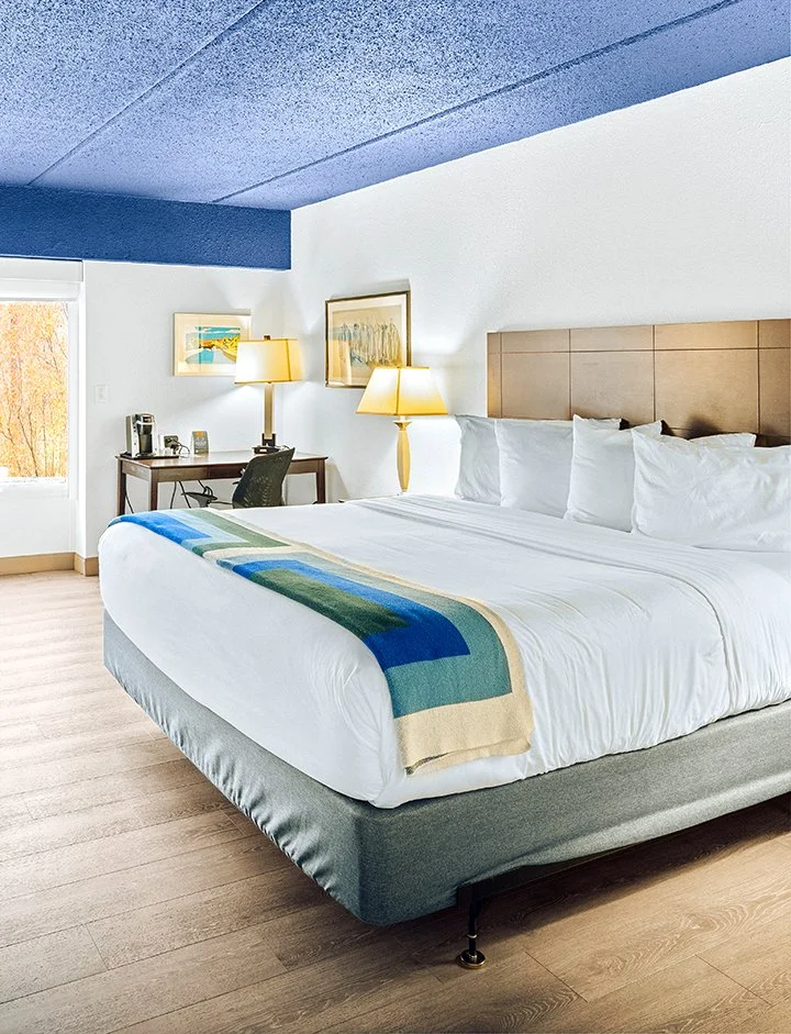 A neatly made hotel room with a king-sized bed, featuring white bedding and pillows, a beige headboard, a colorful blanket at the foot, a wooden desk with a chair, lamps, a coffee maker, and framed pictures on the wall. There is a window showing trees outside.