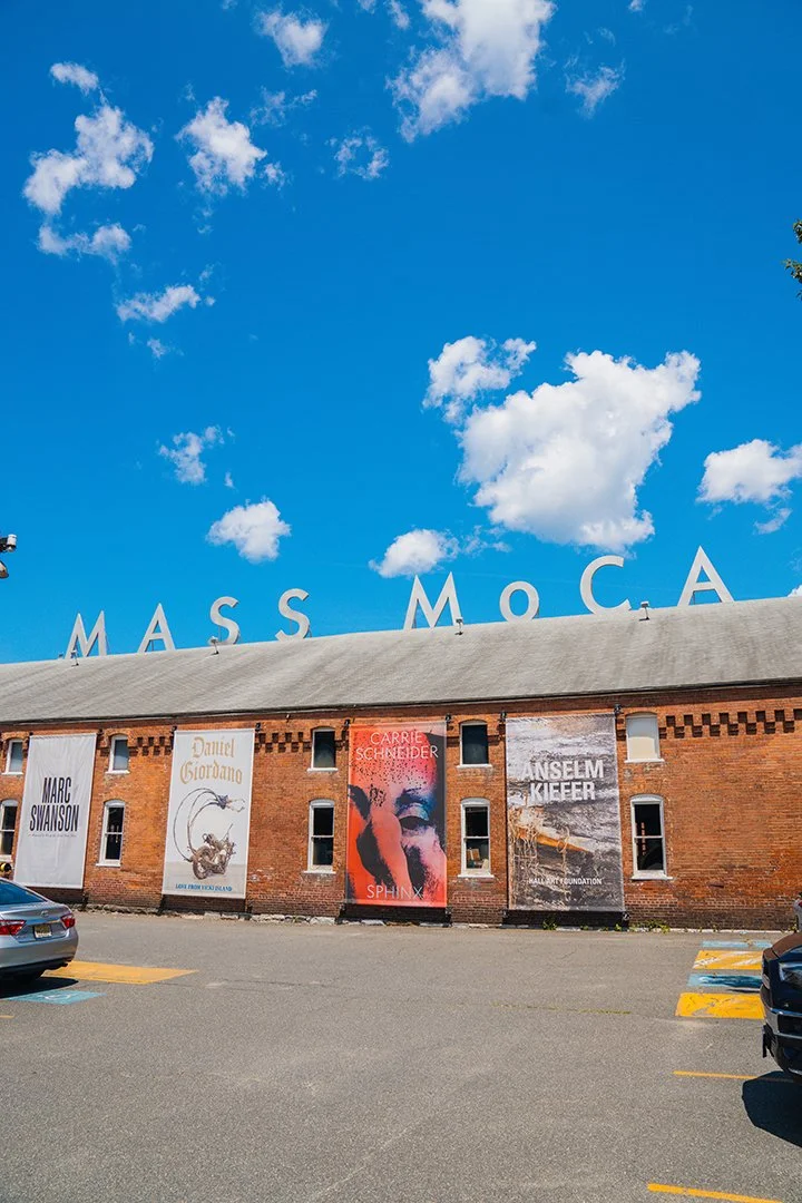 Outdoor view of a brick building with large white letters spelling 'MASS MOCA' on the roof, and four vertical banners on the front showcasing artwork or artists, with a parking lot in the foreground and a blue sky with scattered clouds overhead.