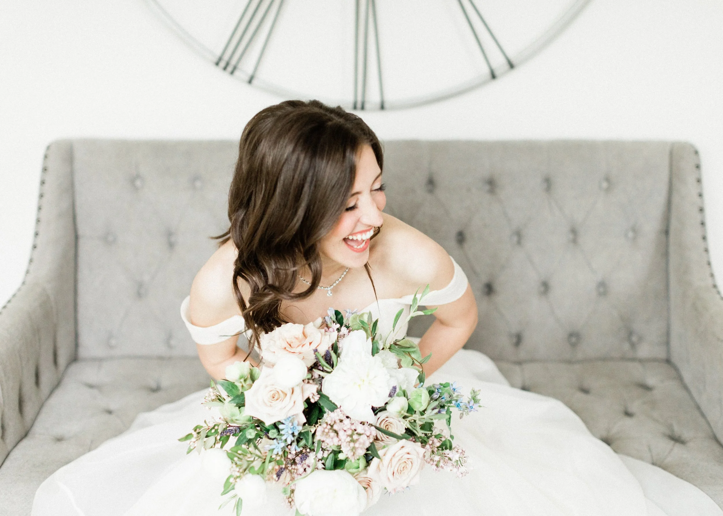 Group of women dressed in light blue and white, smiling and holding bouquets of white and purple flowers, at an outdoor wedding.