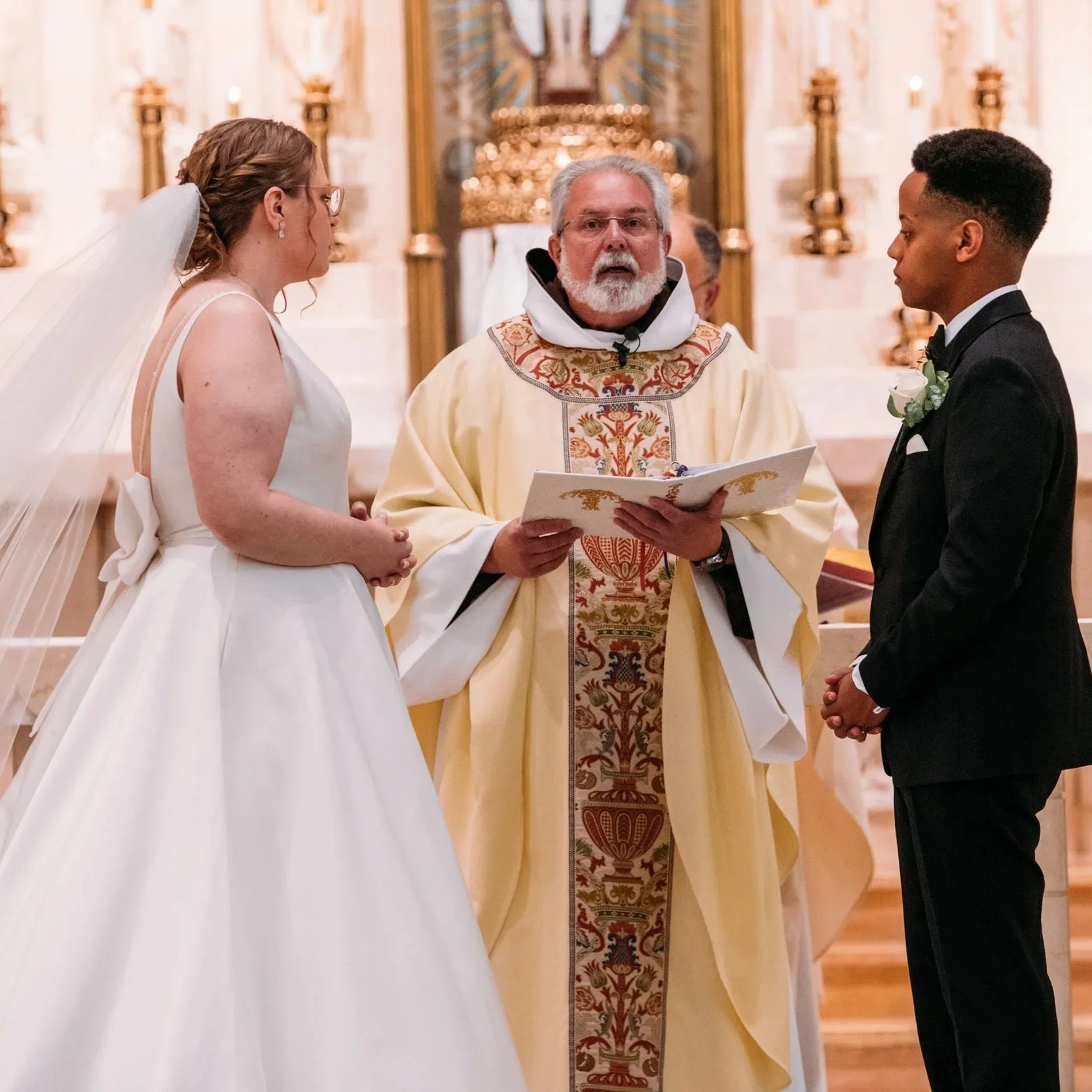 A couple gets married in a church with a priest officiating. The bride wears a white wedding dress and veil, and the groom wears a black tuxedo with a boutonniere. They stand facing each other, holding hands, during the wedding ceremony.