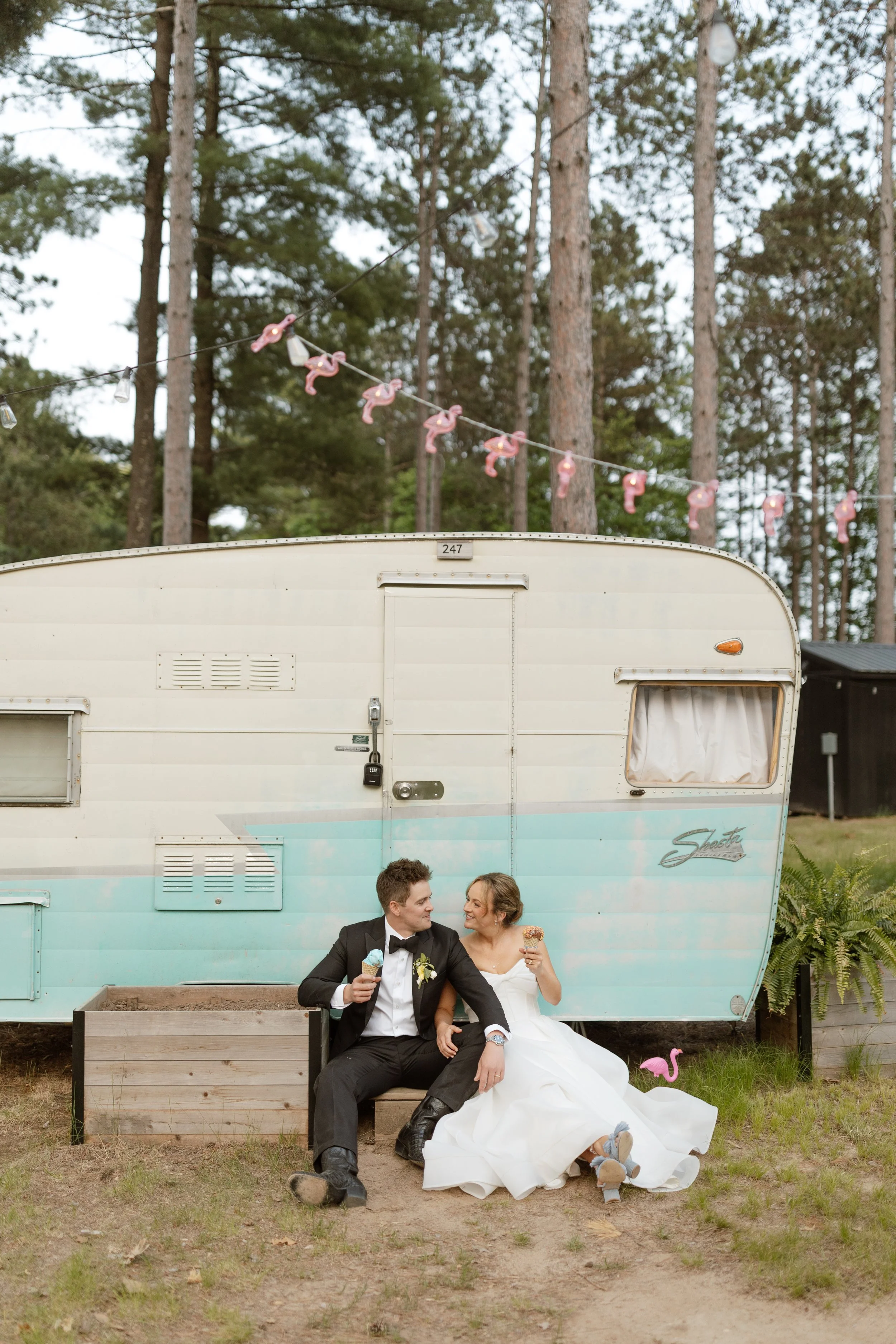 A newlywed couple sitting in front of a vintage camper trailer, sharing ice cream cones and smiling at each other. The groom is dressed in a tuxedo, and the bride is in a white wedding dress. The scene is outdoors, with tall trees and string lights in the background.