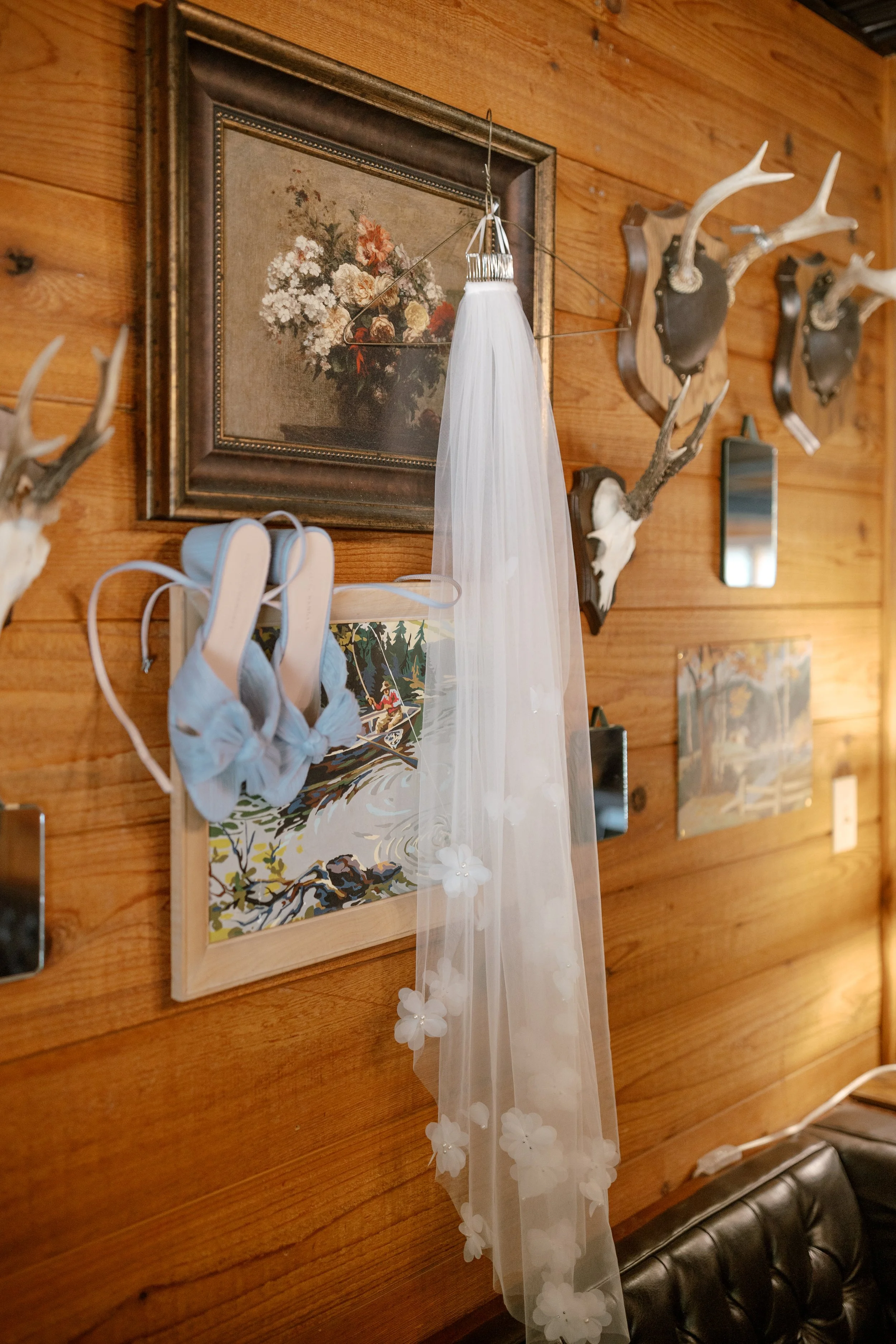 Decorative wall with framed floral painting, mounted deer antlers, a mirror, and a wedding dress veil hanging from a hanger, in a room with wooden walls.